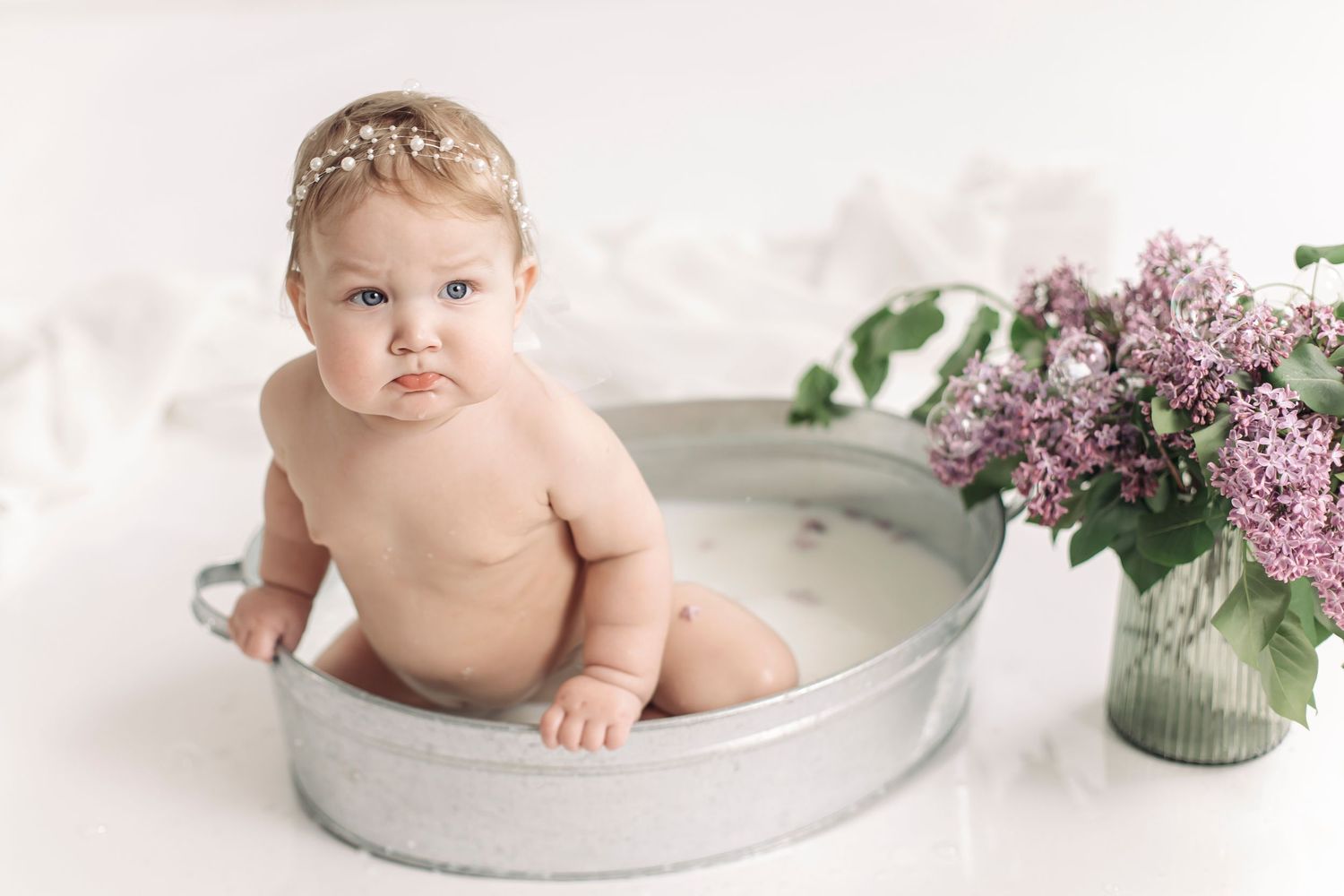 Adorable baby girl sitting in a vintage metal tub with lilac flowers, wearing a delicate floral headband.