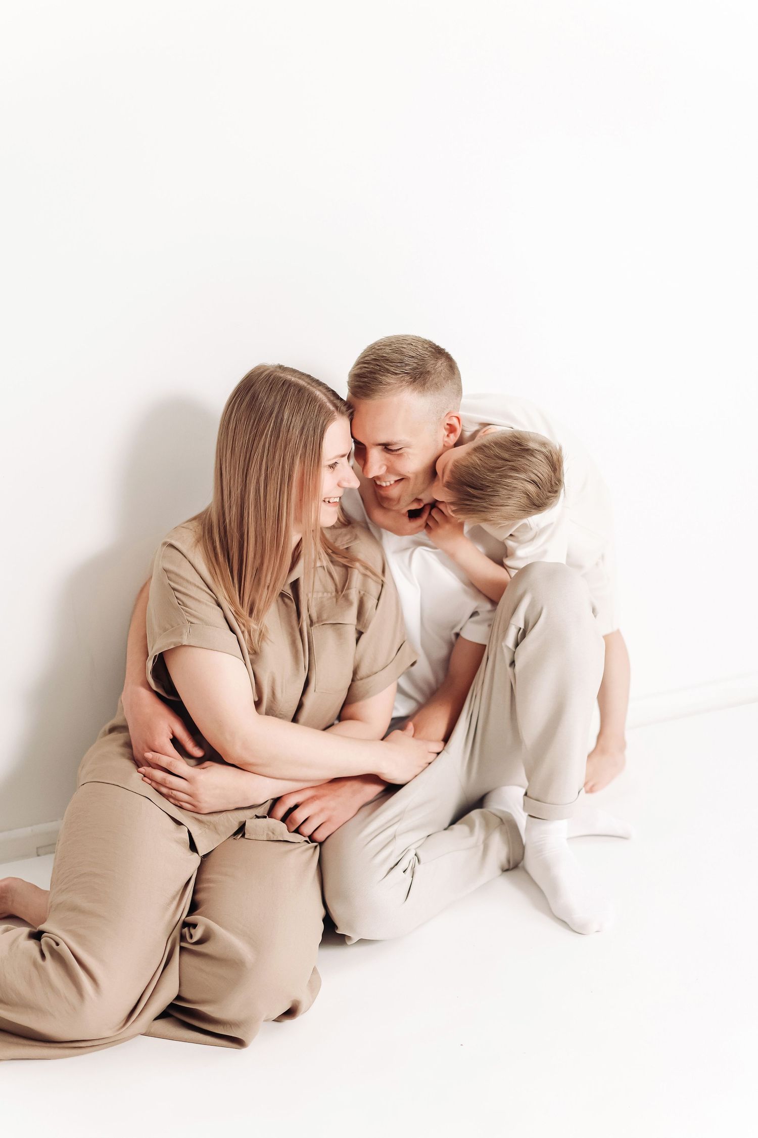 Happy family of three sitting on white floor, parents smiling at each other while child kisses father's cheek.