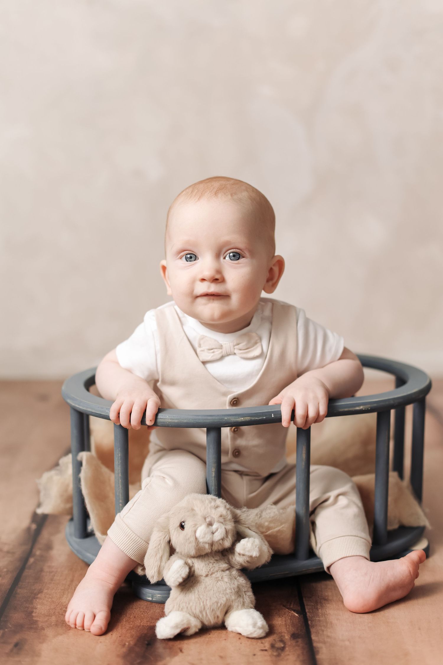 Adorable baby in beige bow tie outfit sits in wooden crib with stuffed bunny toy on hardwood floor.