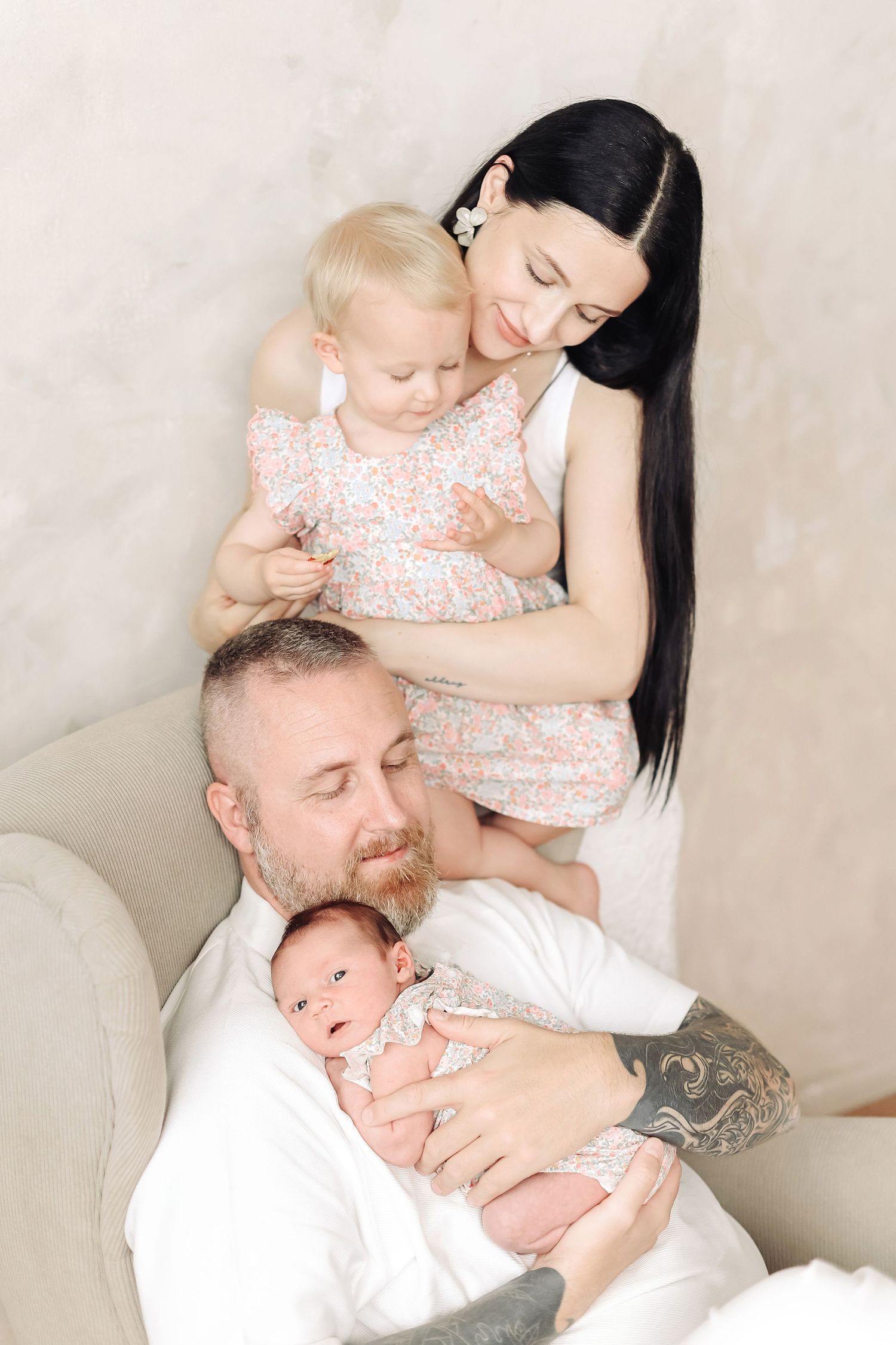 A tattooed father holds a newborn while a dark-haired mother embraces a blonde toddler in matching floral dresses.