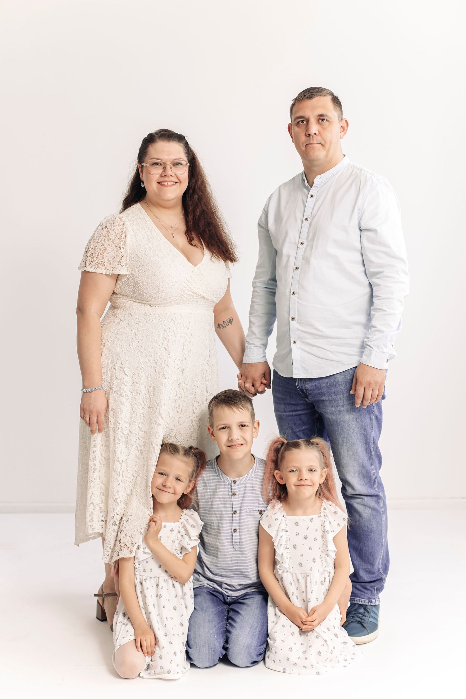 A family of five poses in a white studio, parents holding hands behind three young children in matching outfits.