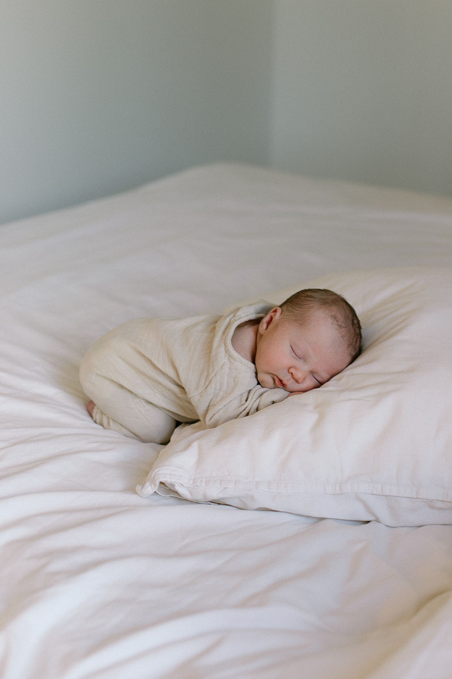 Newborn baby sleeping peacefully on a white pillow, wrapped in a cream swaddle on a soft white bed.
