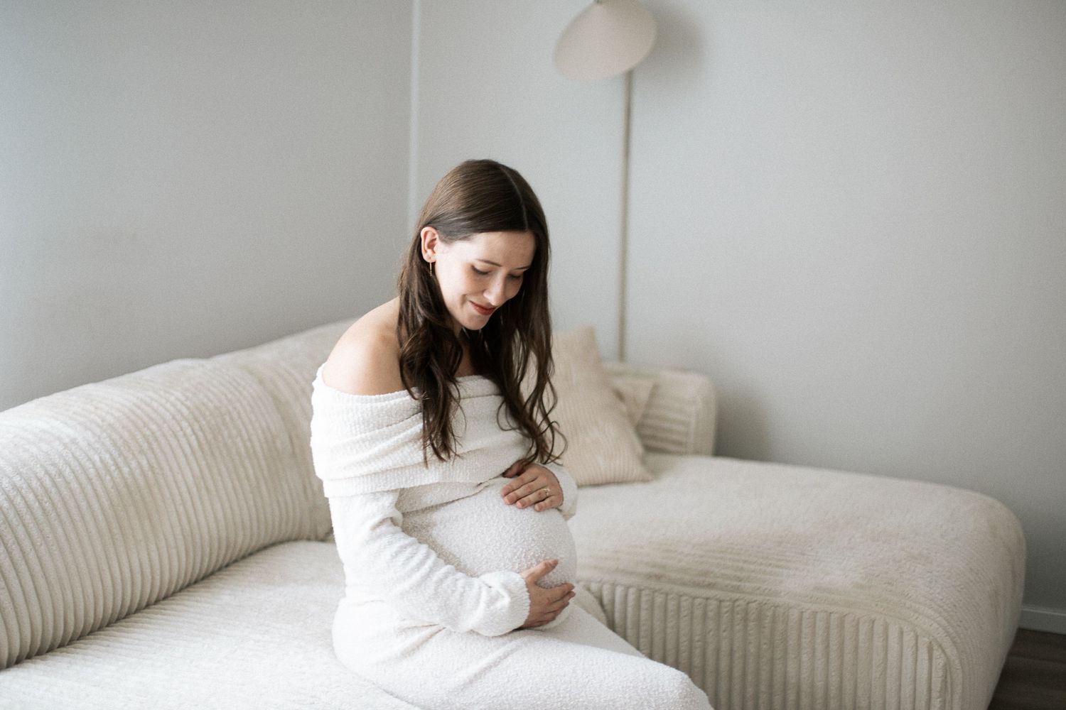 Pregnant woman in white dress sitting on beige sofa, cradling baby bump in bright minimalist room.