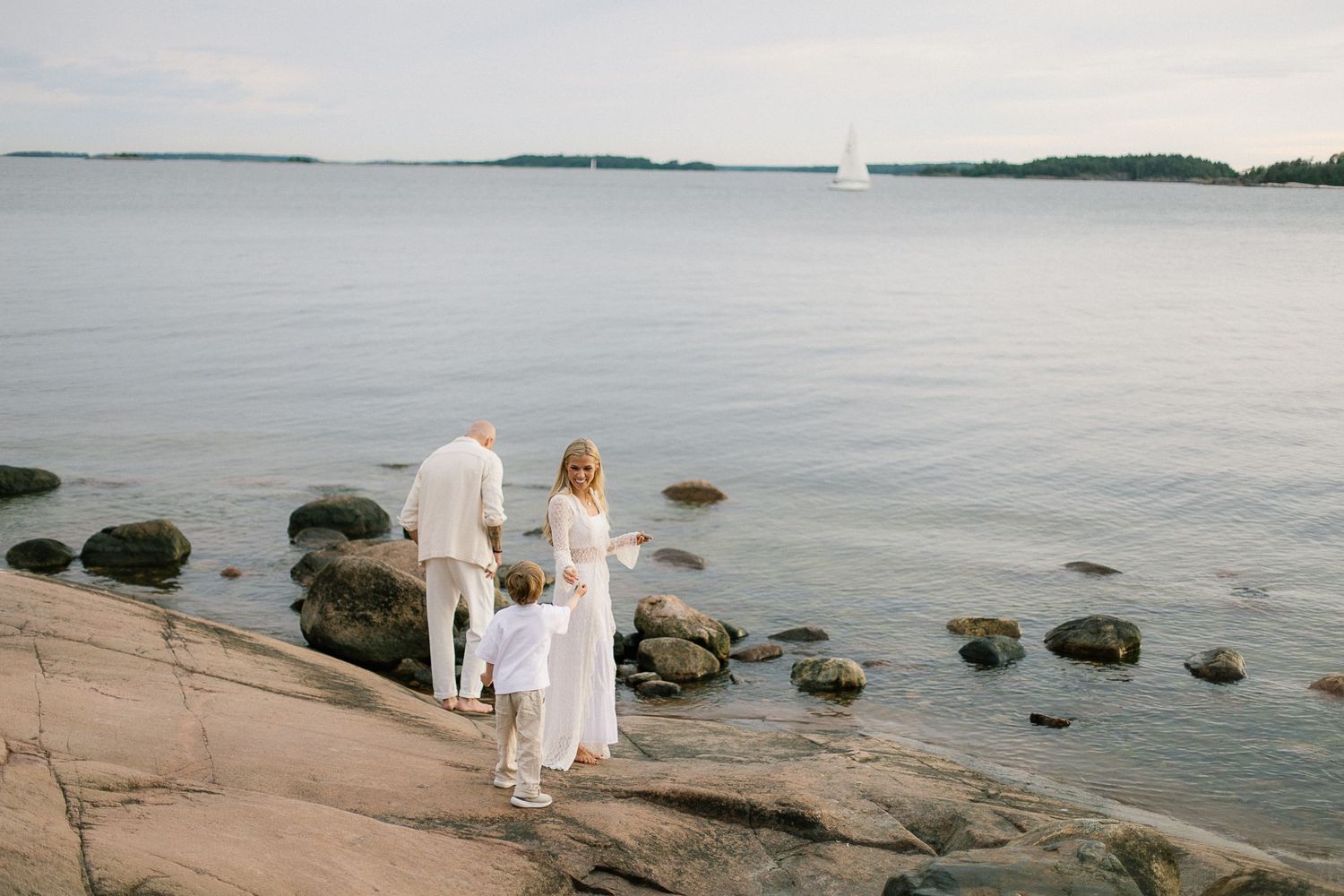 Family of three standing on rocky shoreline in white outfits, gazing at calm sea with sailboat in distance.