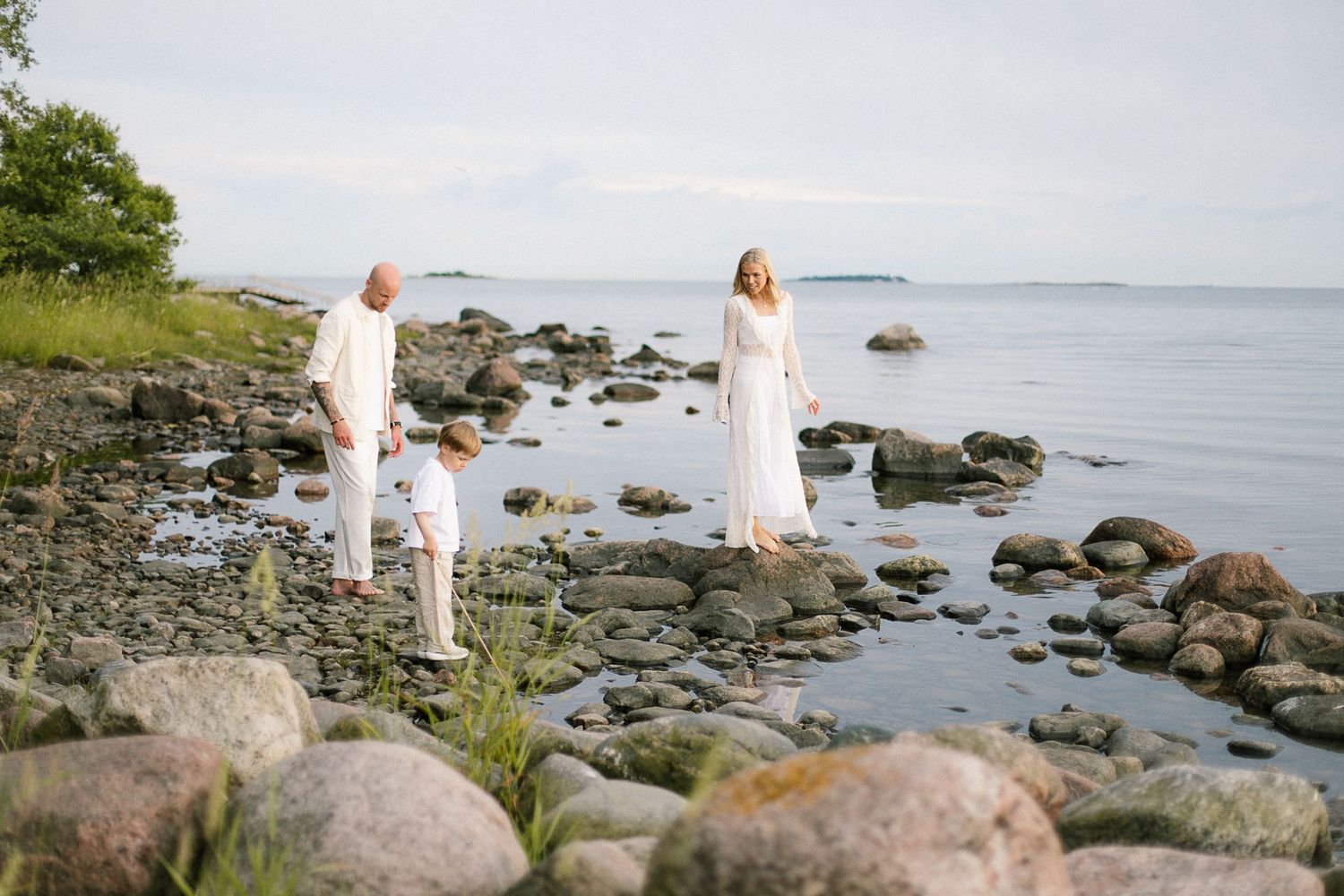 Couple in white outfits standing on rocky shoreline with calm sea and green trees in background.