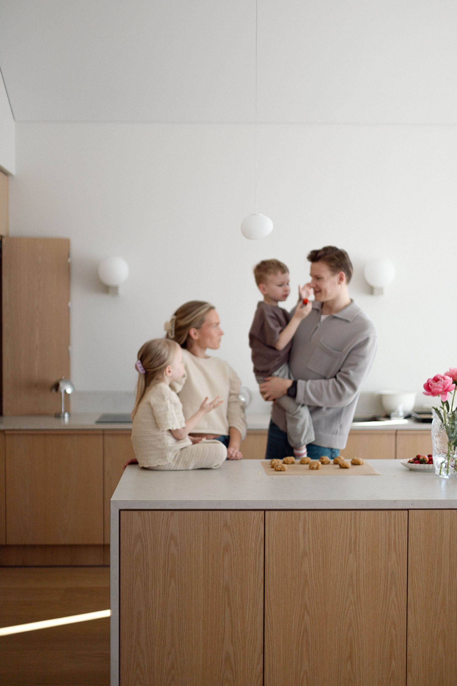 Happy family with two young children sharing a snack in a modern Scandinavian kitchen with warm wood cabinets.