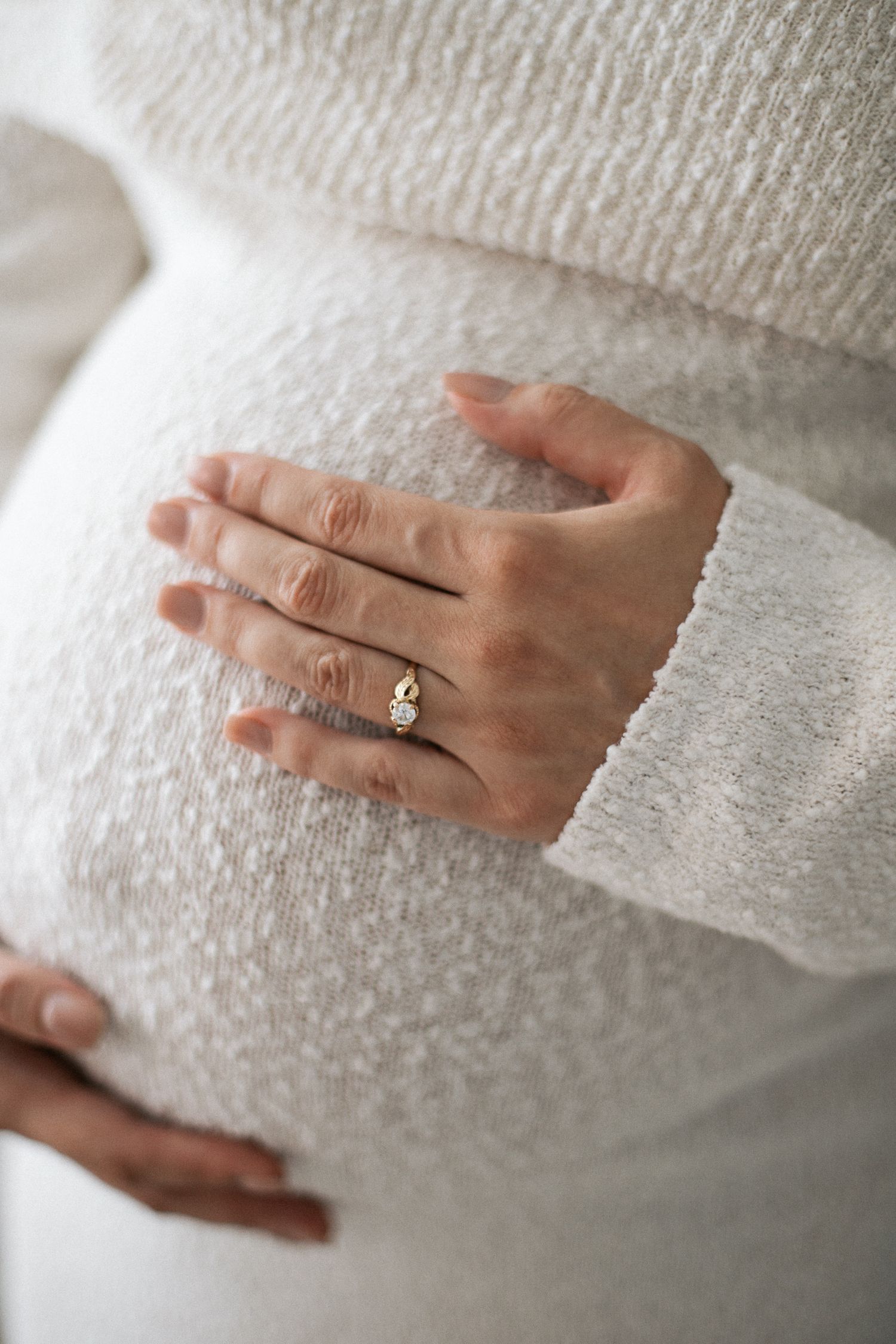Pregnant woman in white sweater gently cradling baby bump, wearing gold ring, close-up maternity photo.
