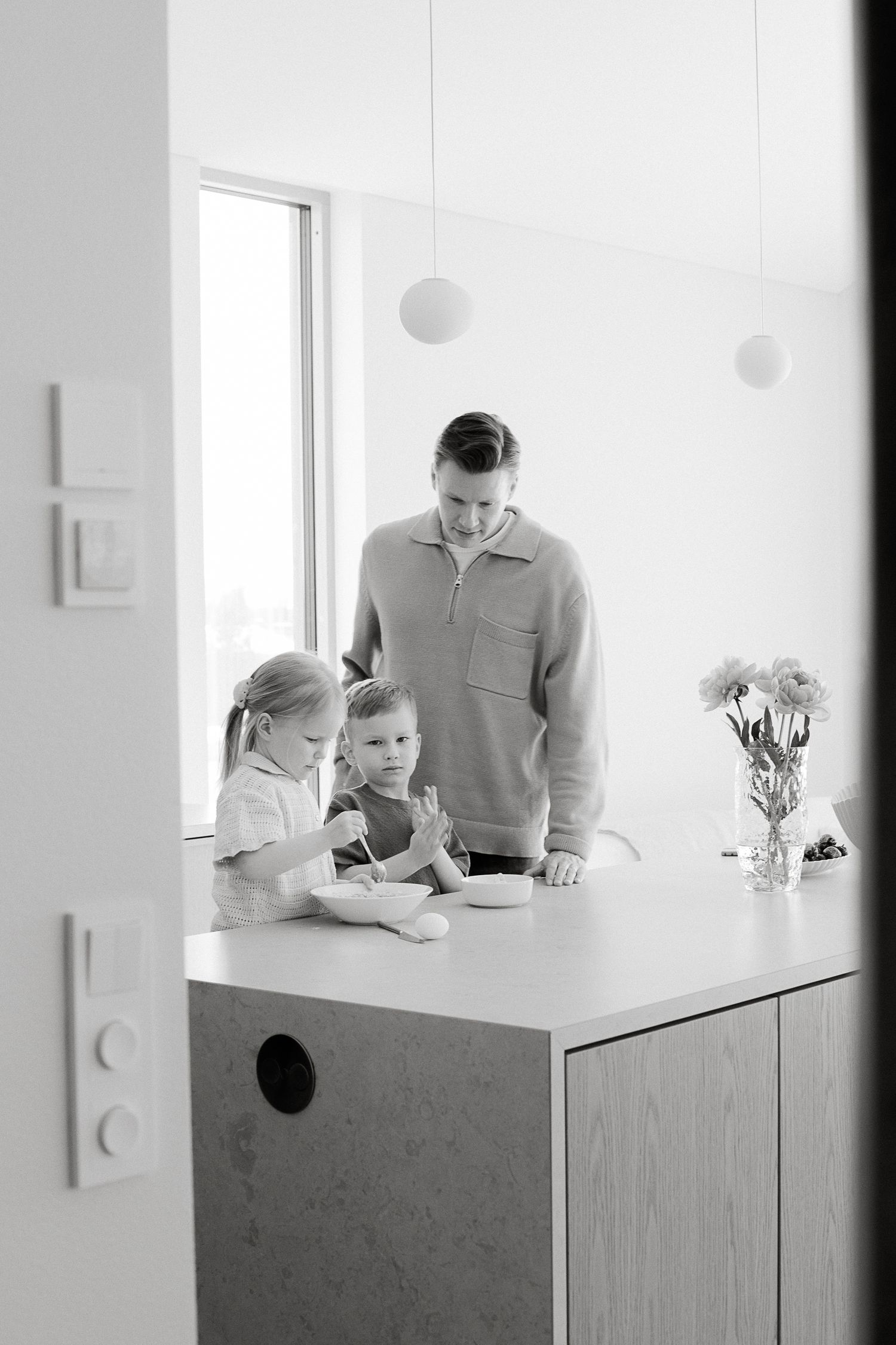 Black and white photo of a father watching his two young children eat at a modern kitchen island.