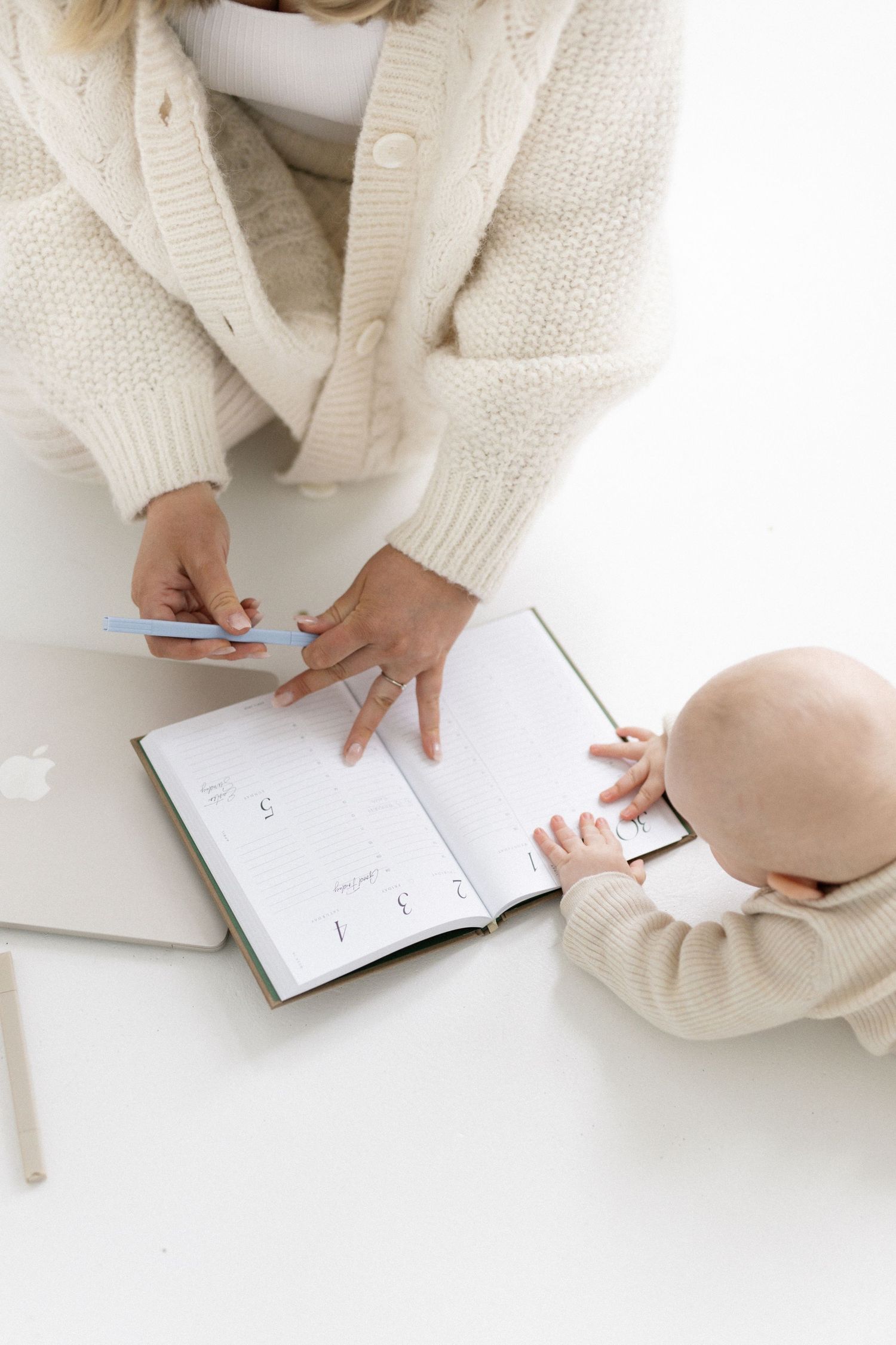 Mother in cream knit cardigan planning with open planner and baby at white desk with laptop.