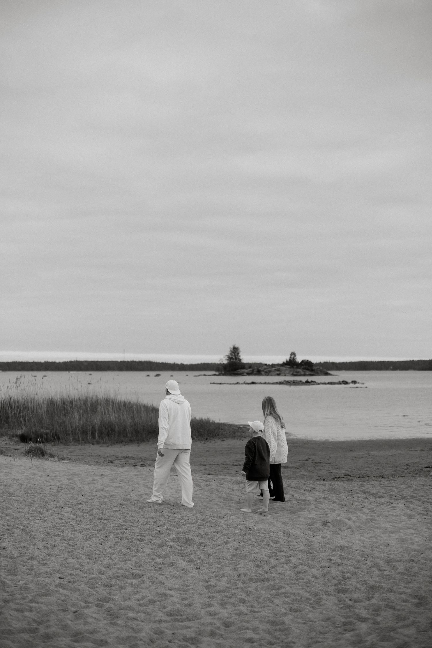 Black and white photo of two people walking on a sandy beach toward calm water under an overcast sky.
