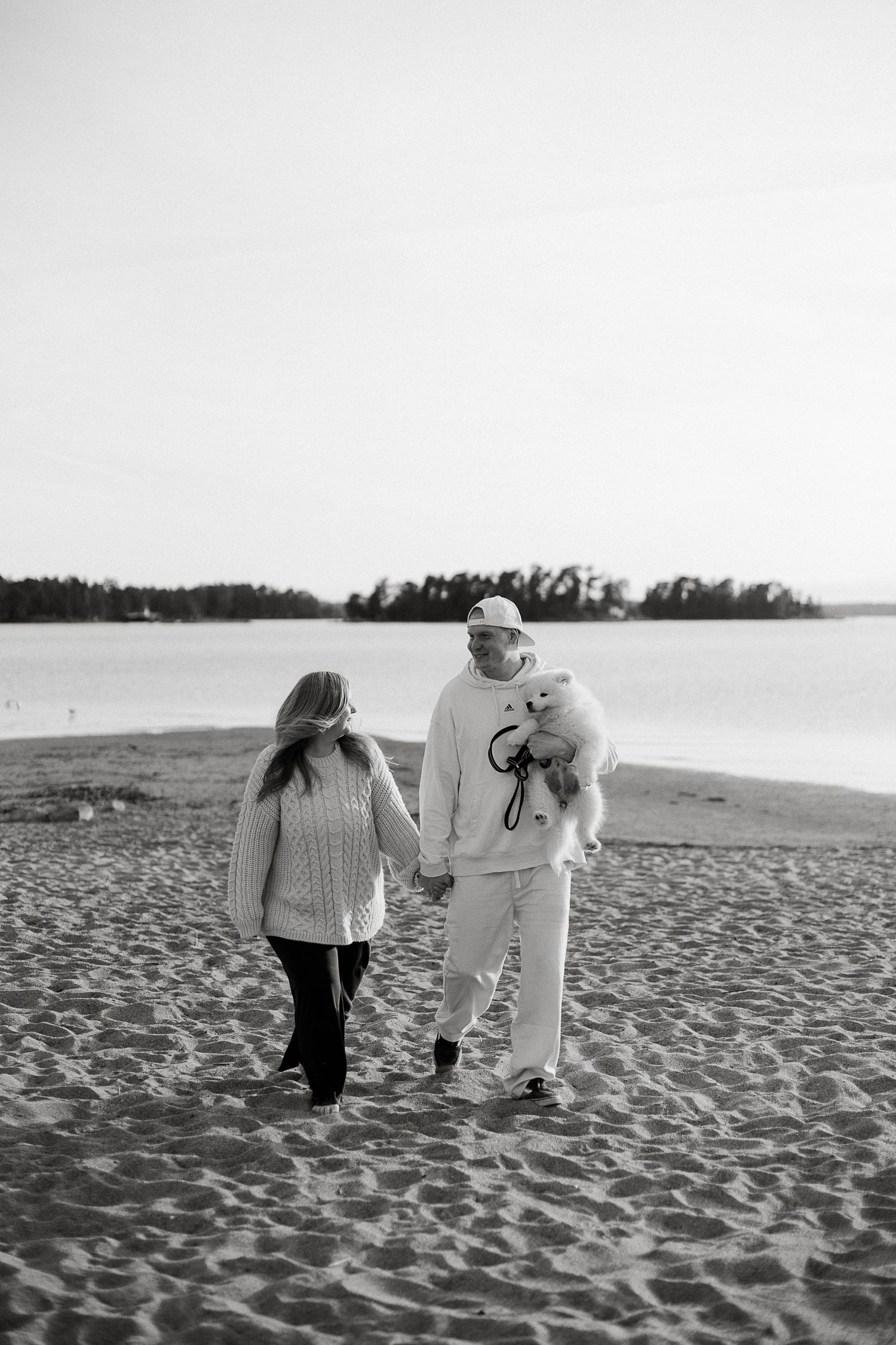 Couple holding hands walking on beach with small white dog, black and white photo.