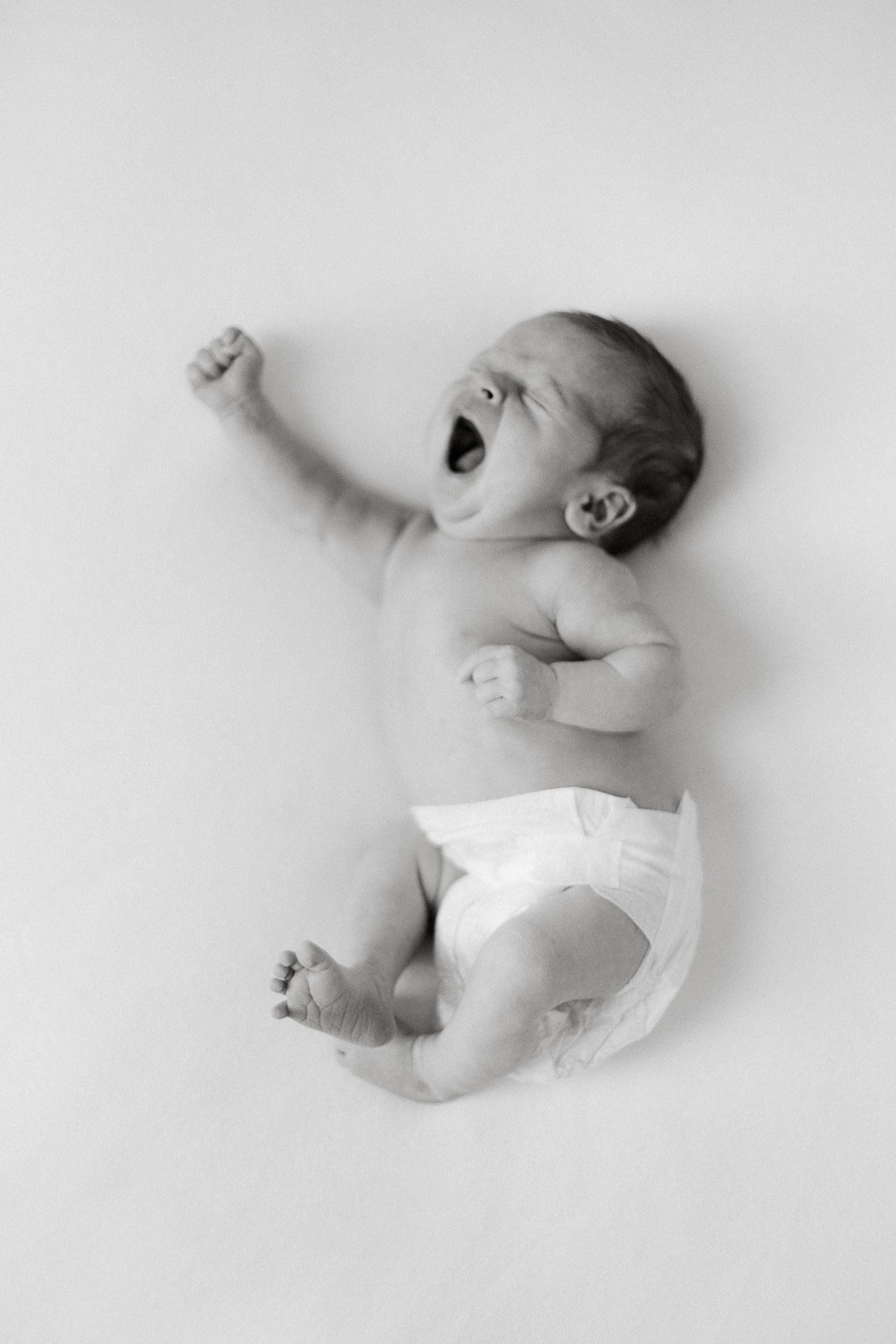 Black and white photo of a yawning newborn baby in a diaper lying on a white background.