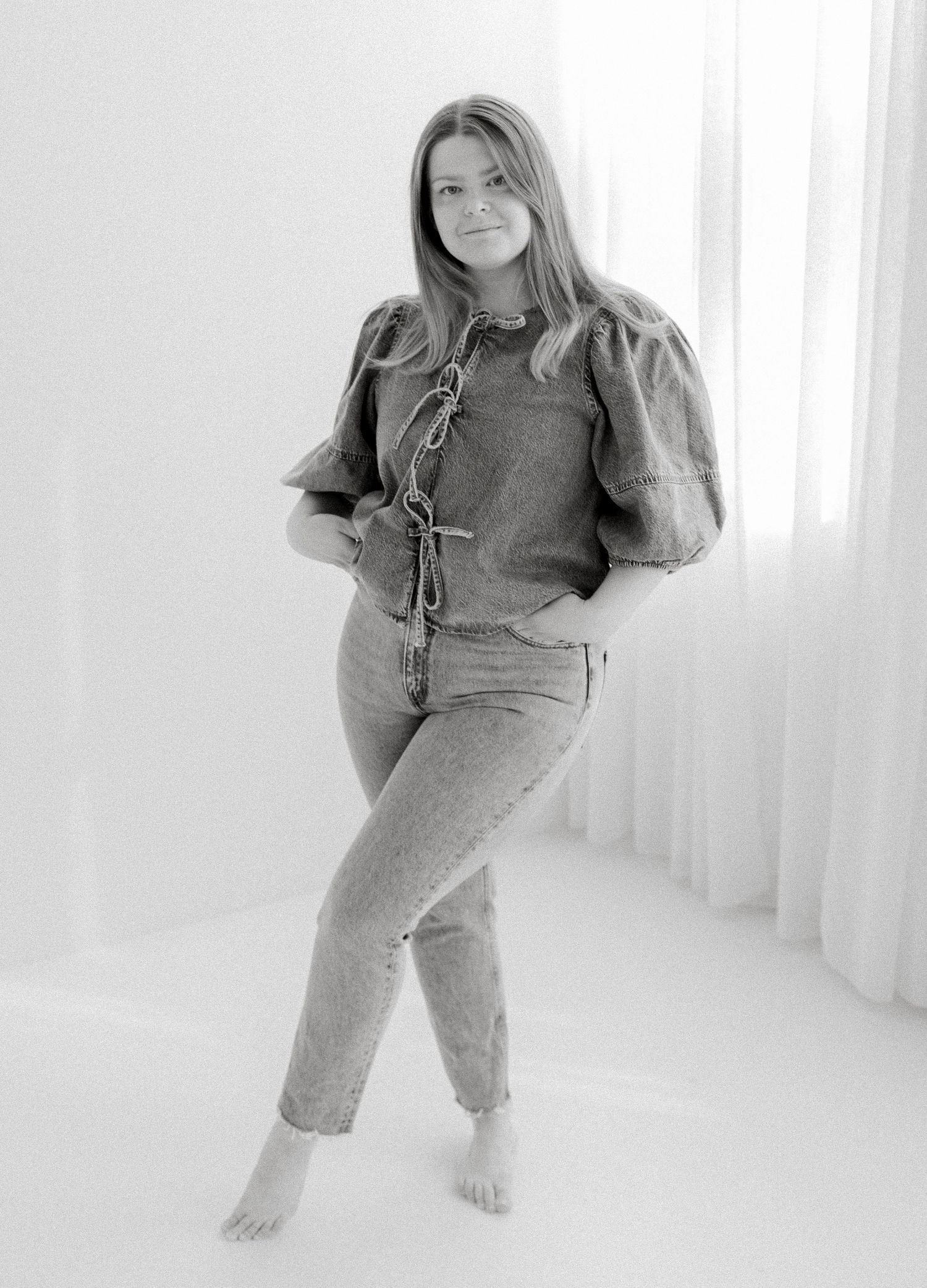 Young woman in jeans and puff-sleeve top poses barefoot in bright white studio, black and white photo.