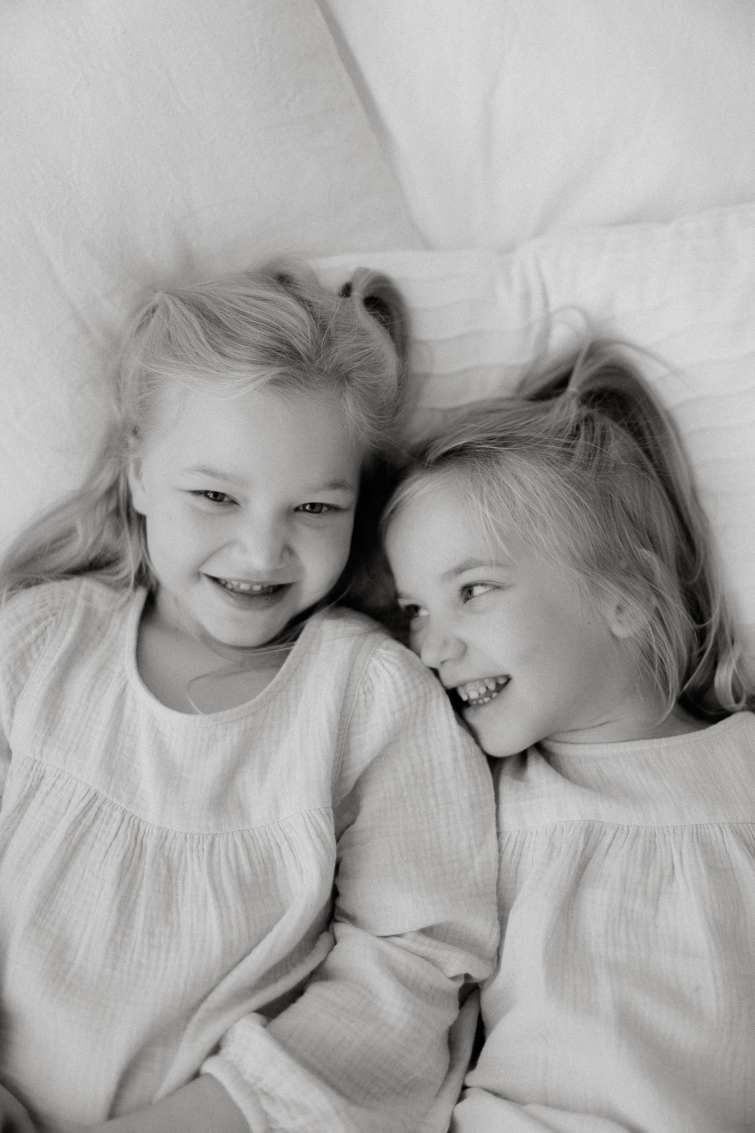 Two joyful blonde sisters in white dresses cuddling and laughing together on white bedding, monochrome photo.