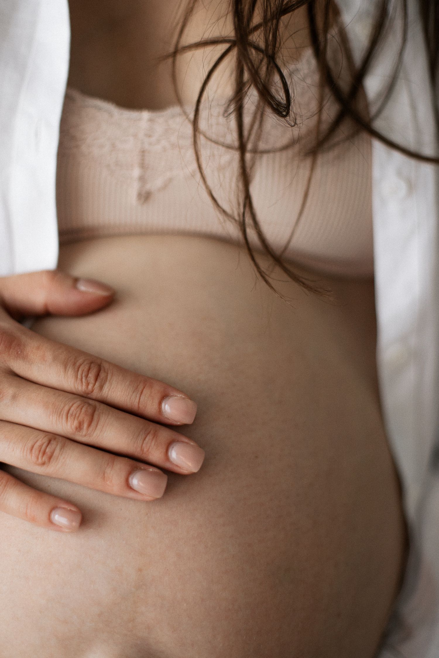 Pregnant woman in beige lace top gently cradling her bare baby bump with one hand.