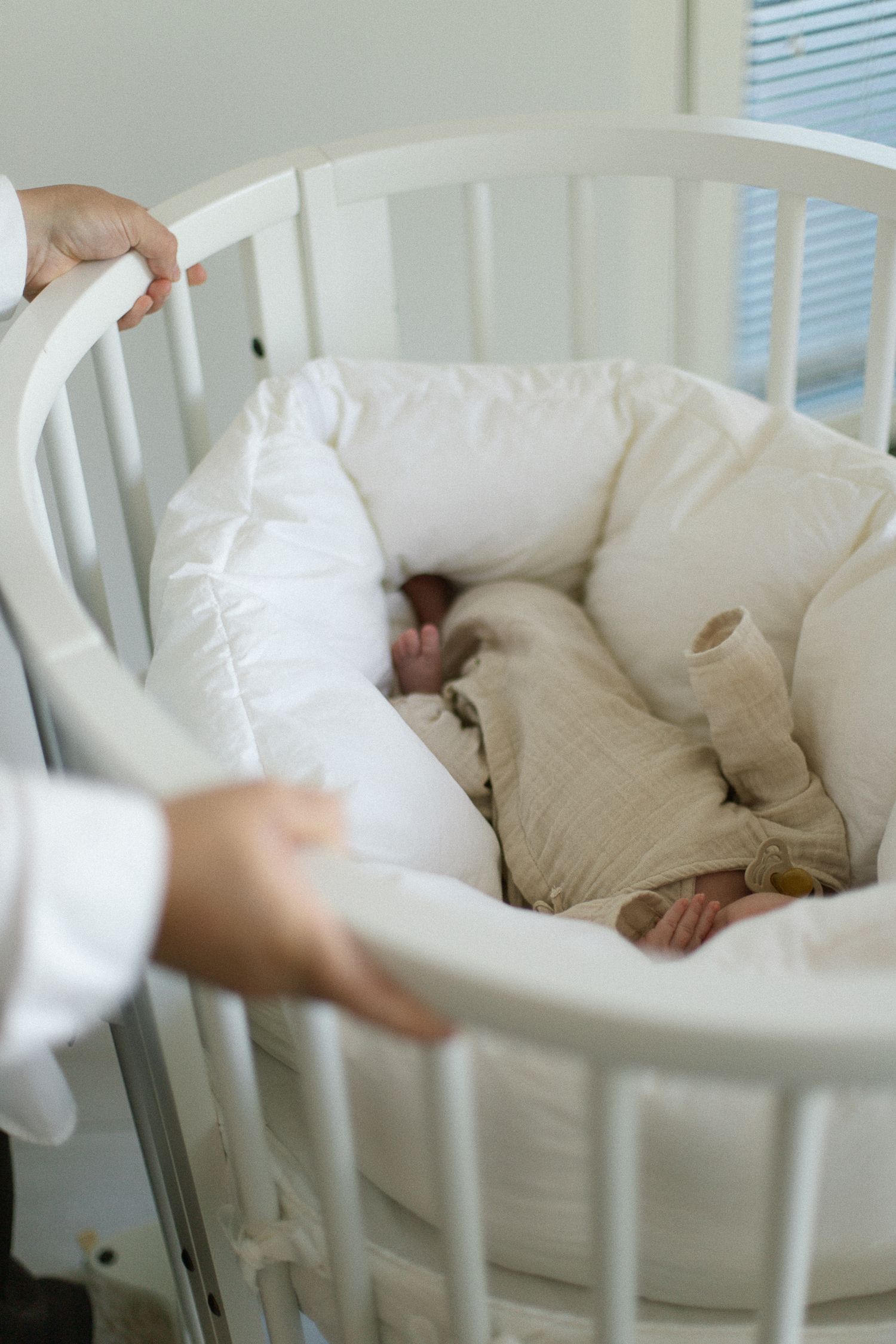 Newborn baby sleeping peacefully in a white oval crib with soft bedding and a beige muslin outfit.