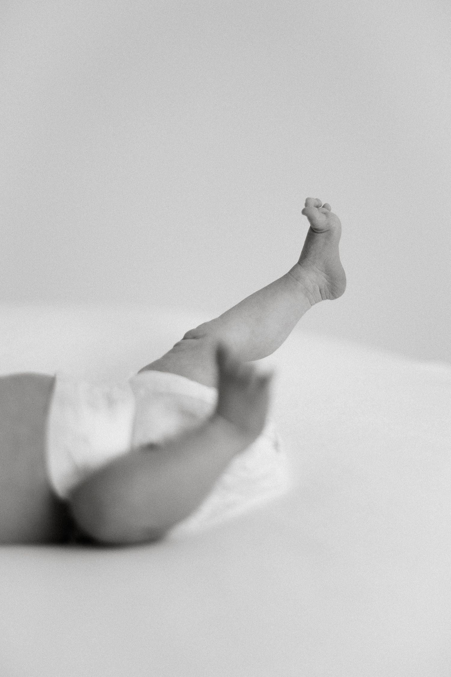 Black and white photo of a baby lying down with one tiny leg raised up, wearing a diaper.