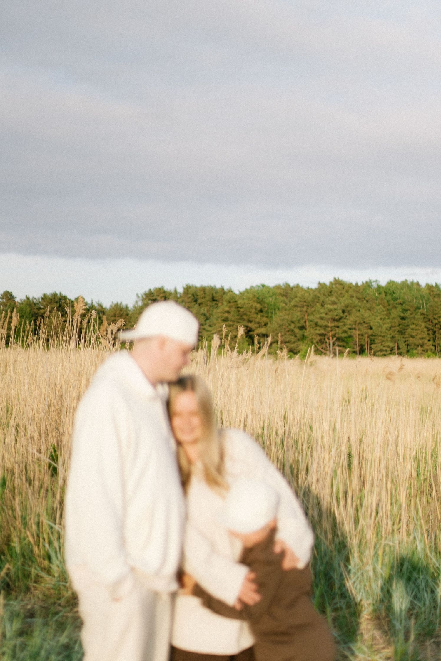 Blurred couple embracing in golden field with evergreen trees and cloudy sky in background.