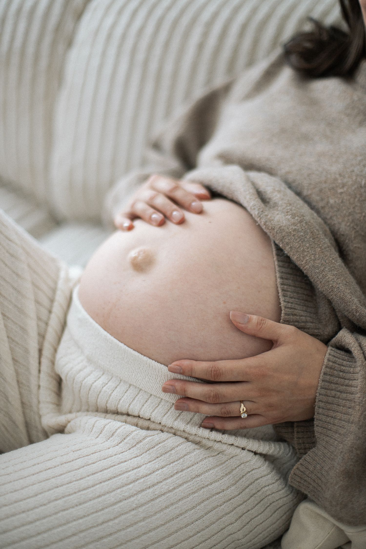Pregnant woman in cream knit sweater cradling her bare belly with both hands while resting.