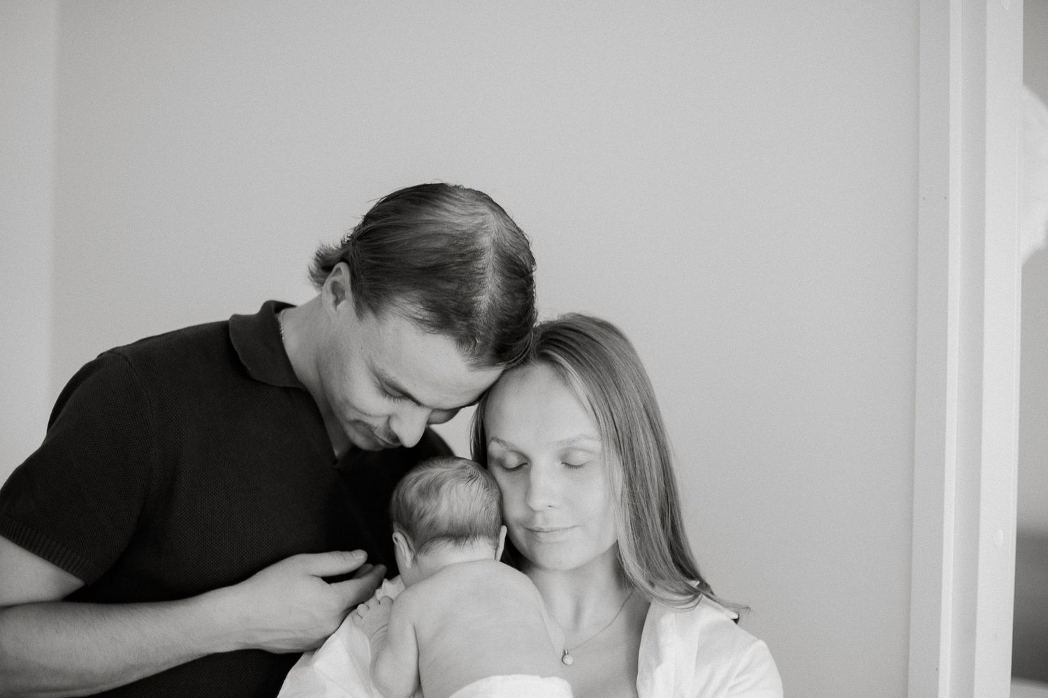 Black and white photo of new parents tenderly cradling their newborn baby, heads bowed together in a loving embrace.