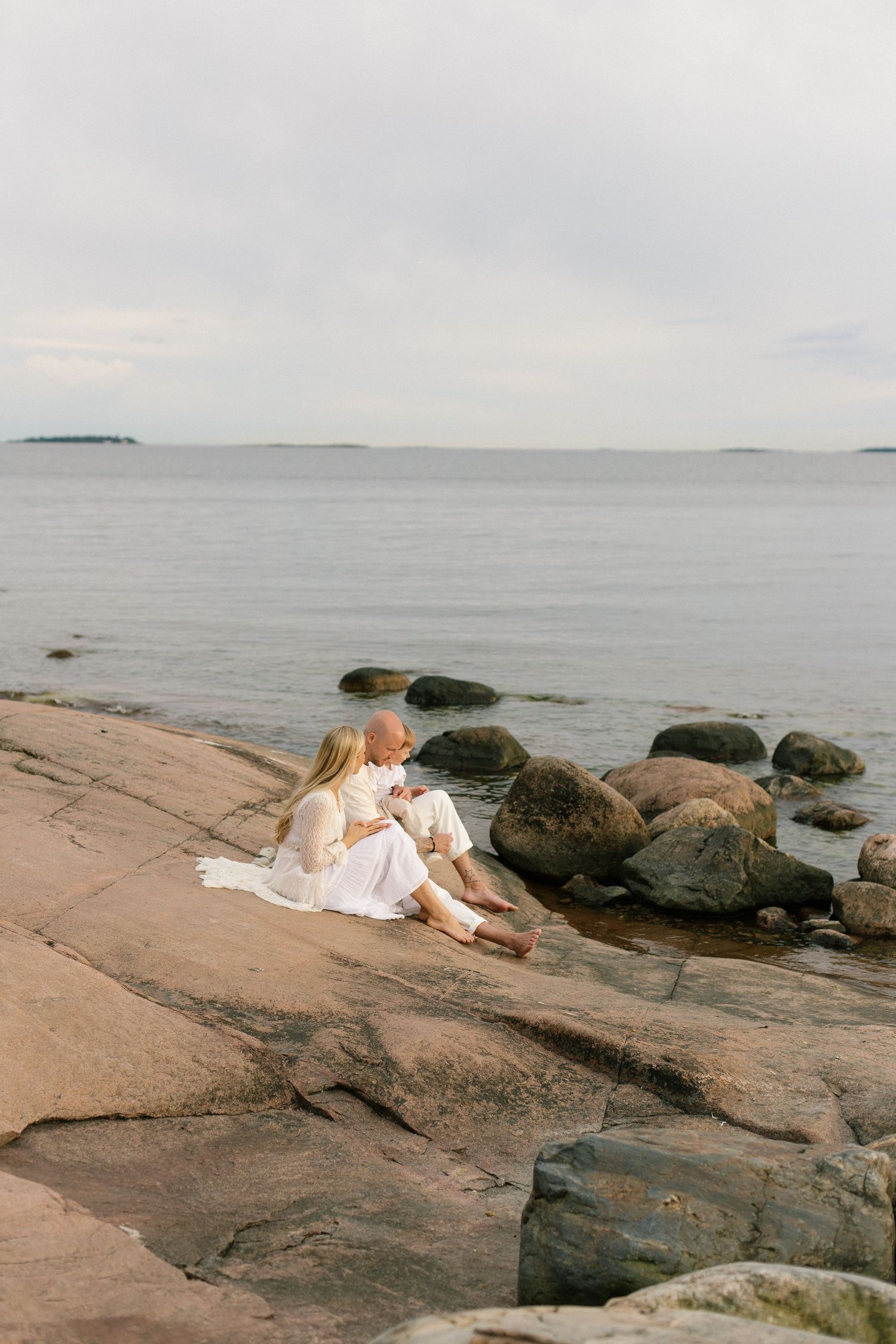 Couple sitting together on rocky shoreline by calm sea, dressed in white, enjoying a peaceful moment outdoors.