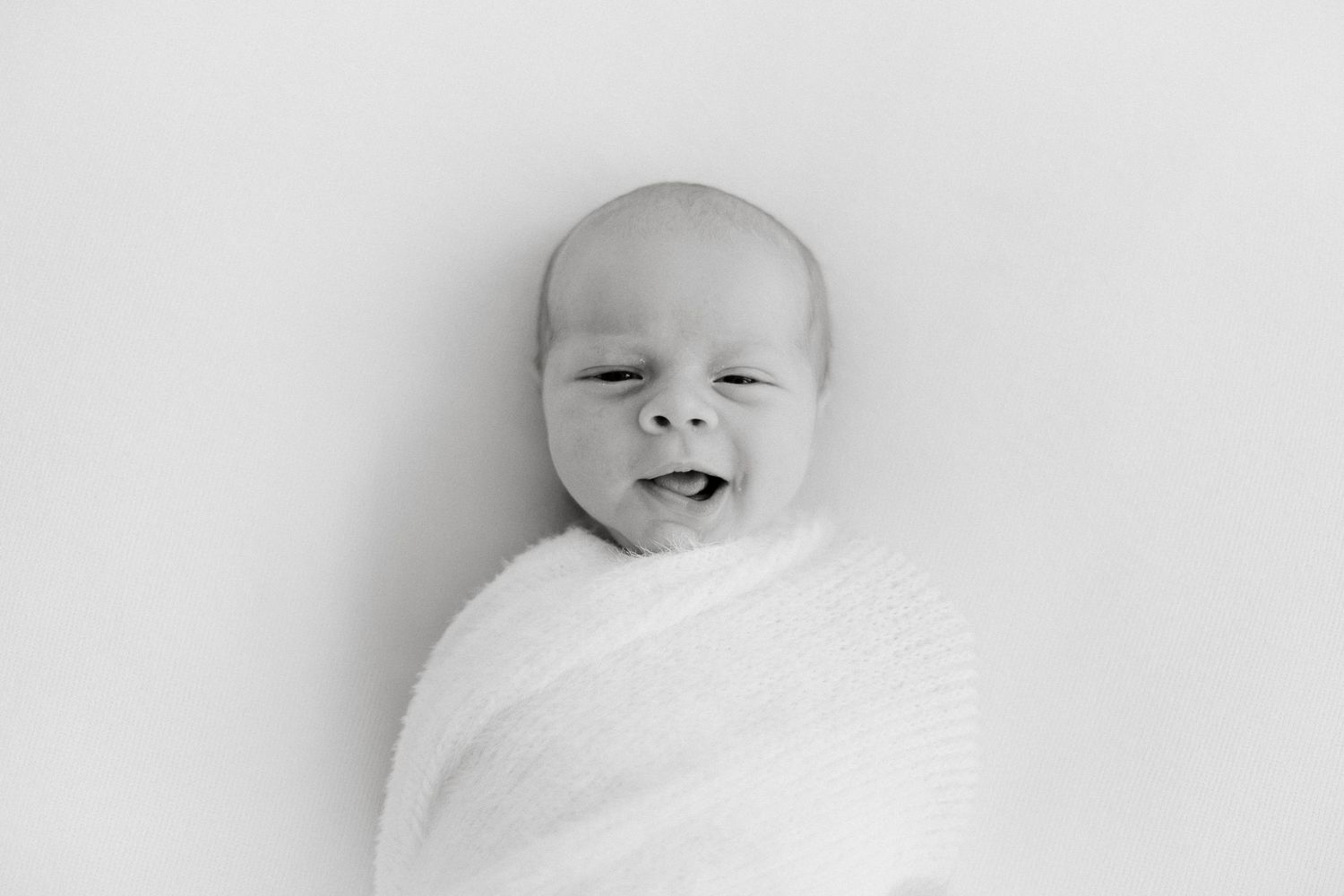 Black and white newborn portrait of a swaddled baby with eyes slightly open, lying on a white background.