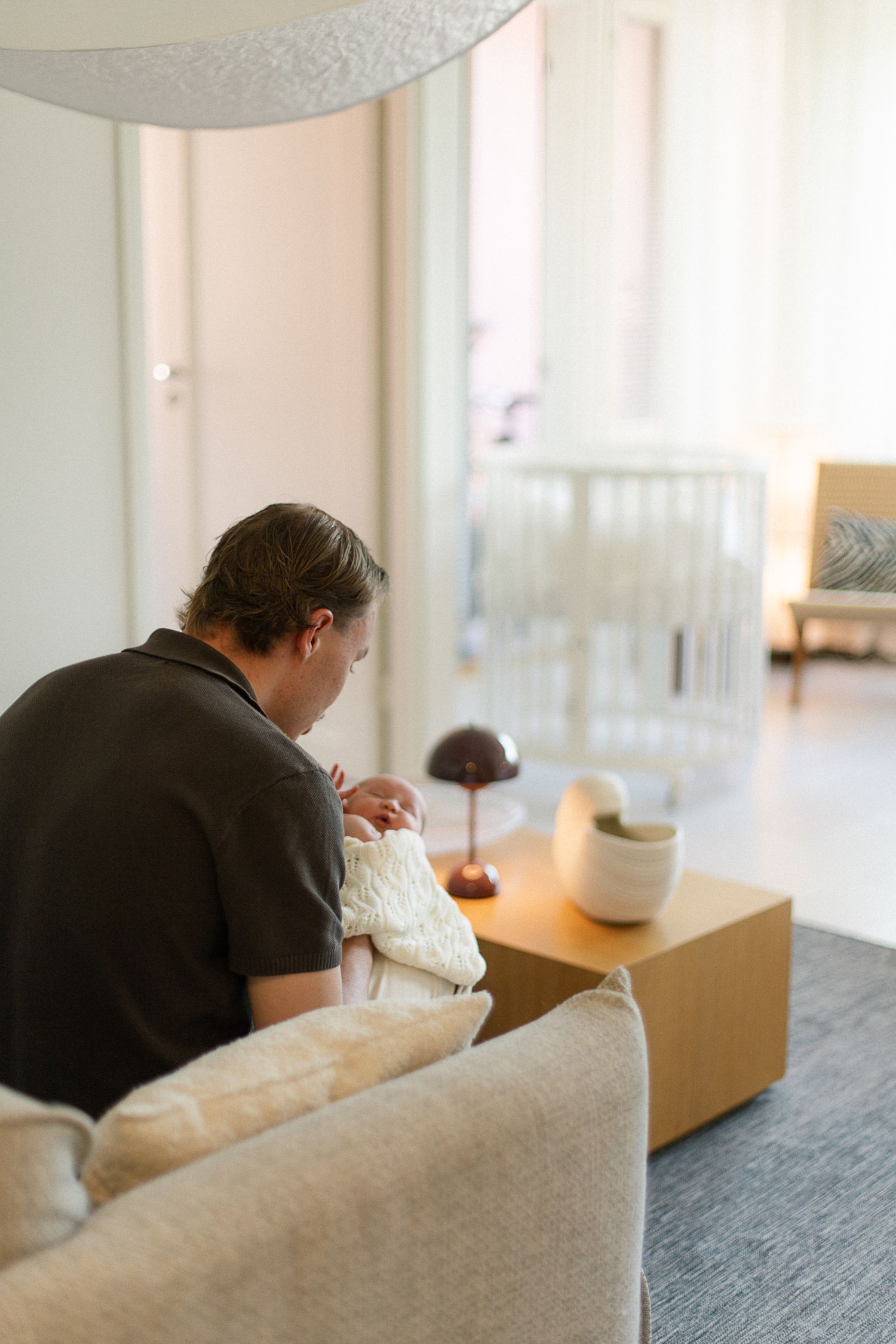 Father sitting on sofa holding newborn baby in white knit blanket, with white crib visible in modern nursery background.