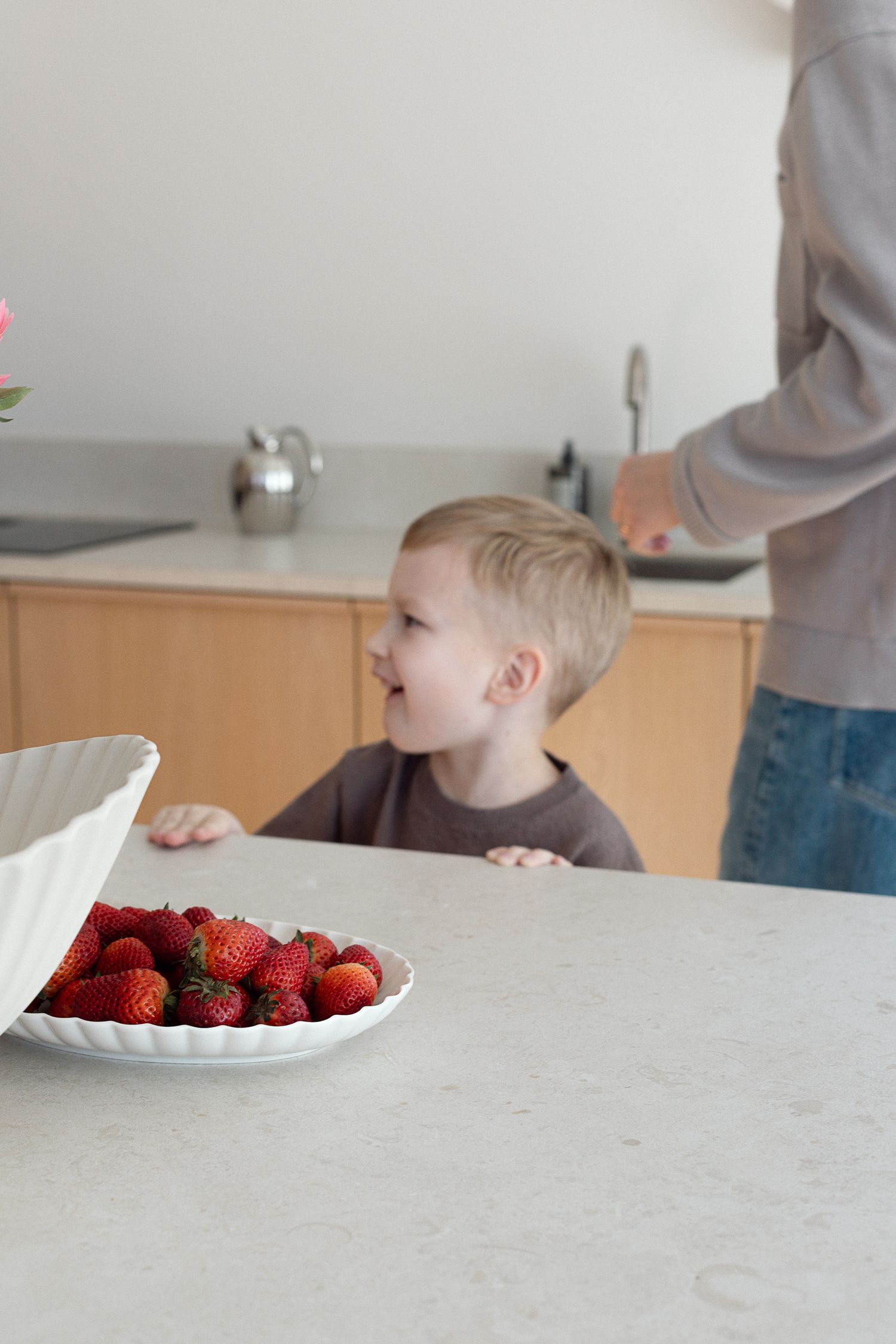 Young blonde toddler smiling at kitchen counter with fresh red strawberries in white bowl.