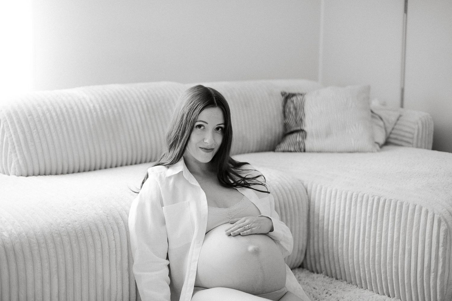 Black and white photo of a pregnant woman sitting on the floor leaning against a white sofa, cradling her baby bump.