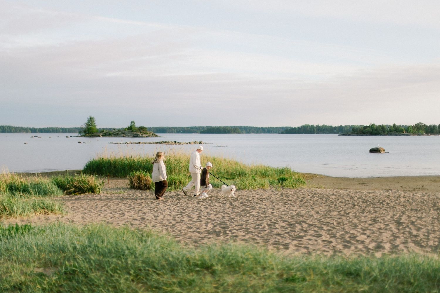 Couple walking white dog on sandy beach by calm lake at dusk, surrounded by green grass and trees.