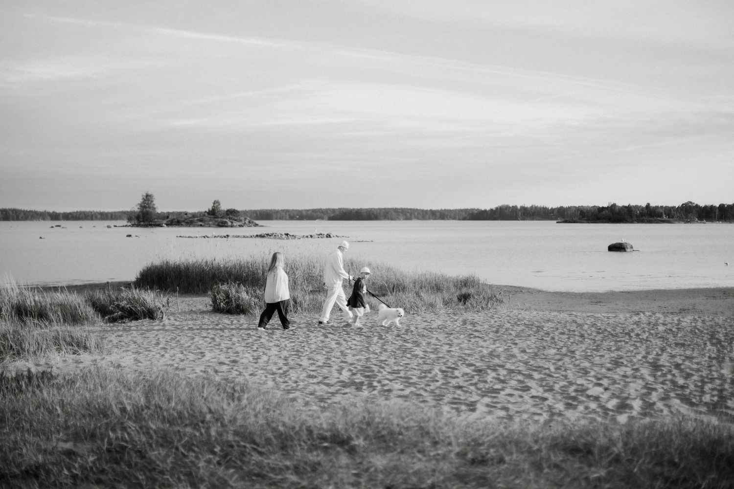 Two people and a dog walking on a sandy beach near calm water in a black and white photo.