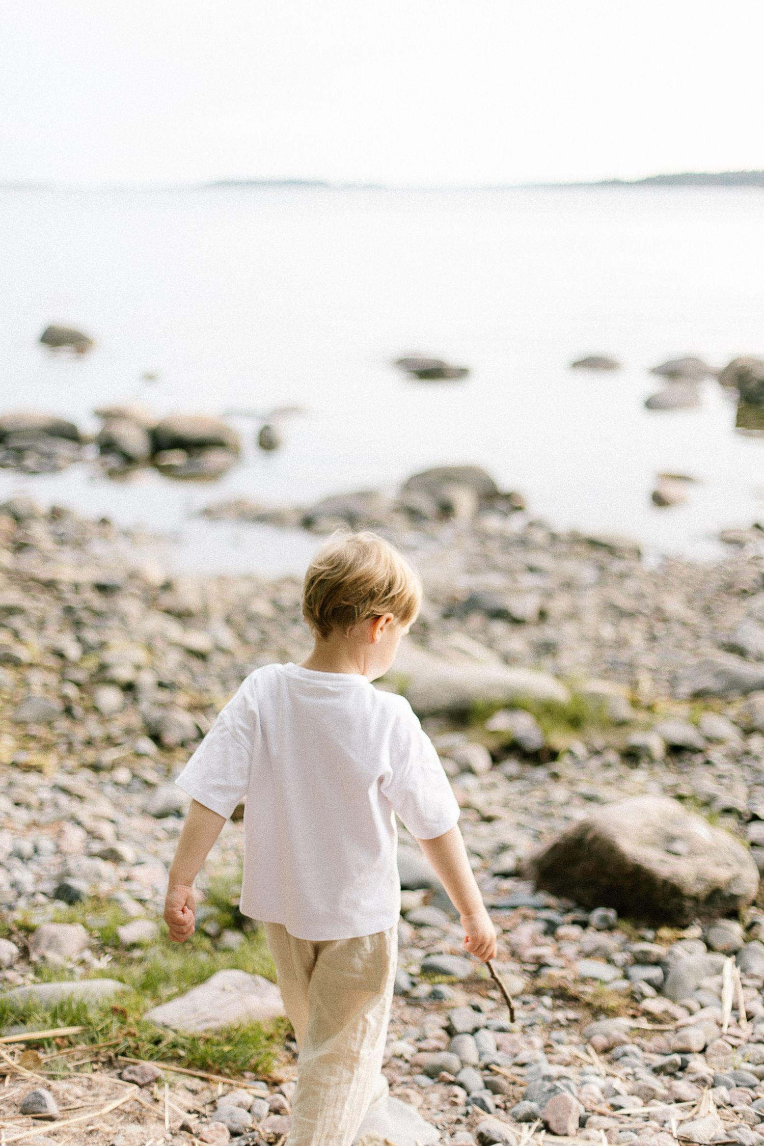 A young boy in a white t-shirt walks along a rocky shoreline, holding a stick while exploring the peaceful waterfront.