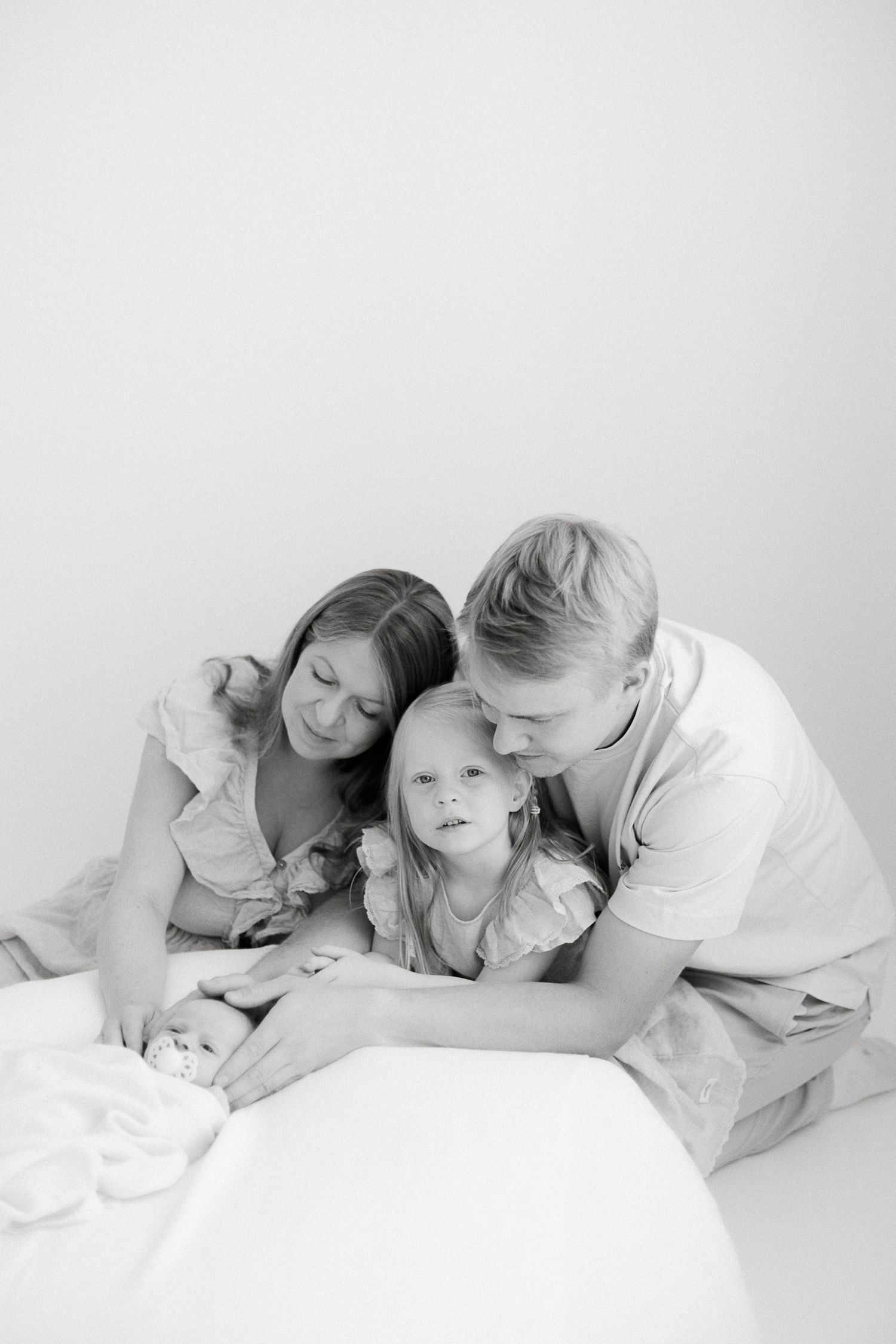 Black and white family photo of parents and toddler girl lovingly gazing at newborn baby on white surface.