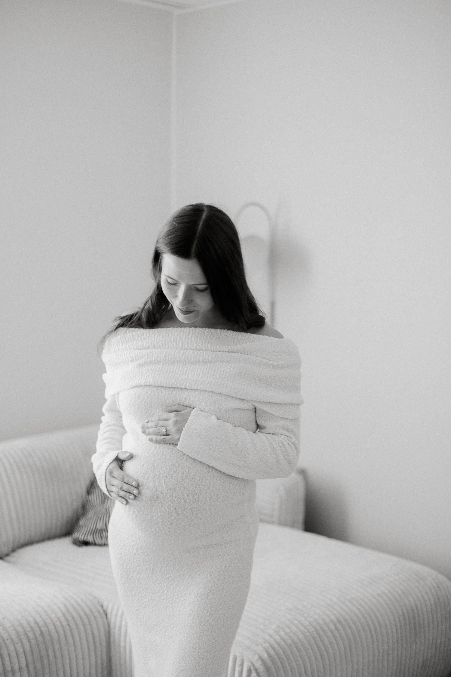 Pregnant woman in white off-shoulder knit dress cradles baby bump in black and white maternity photo.