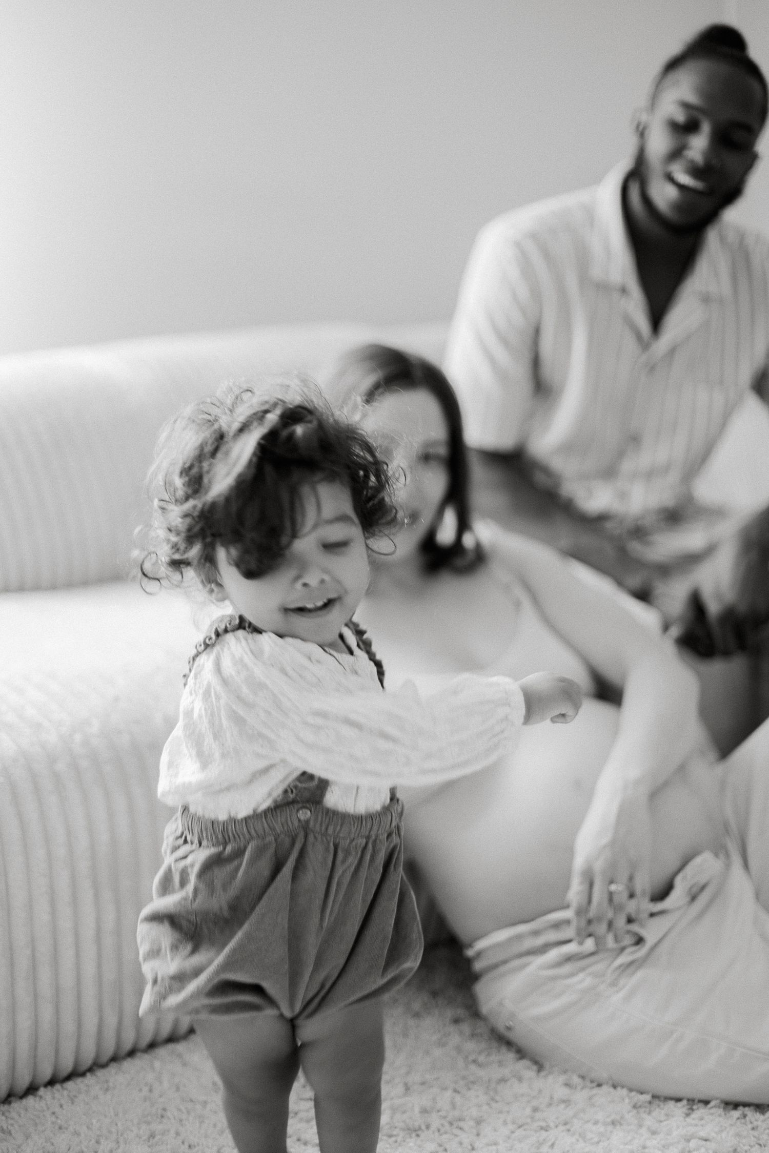 Black and white family maternity photo with toddler touching pregnant mother's belly while father smiles in background.