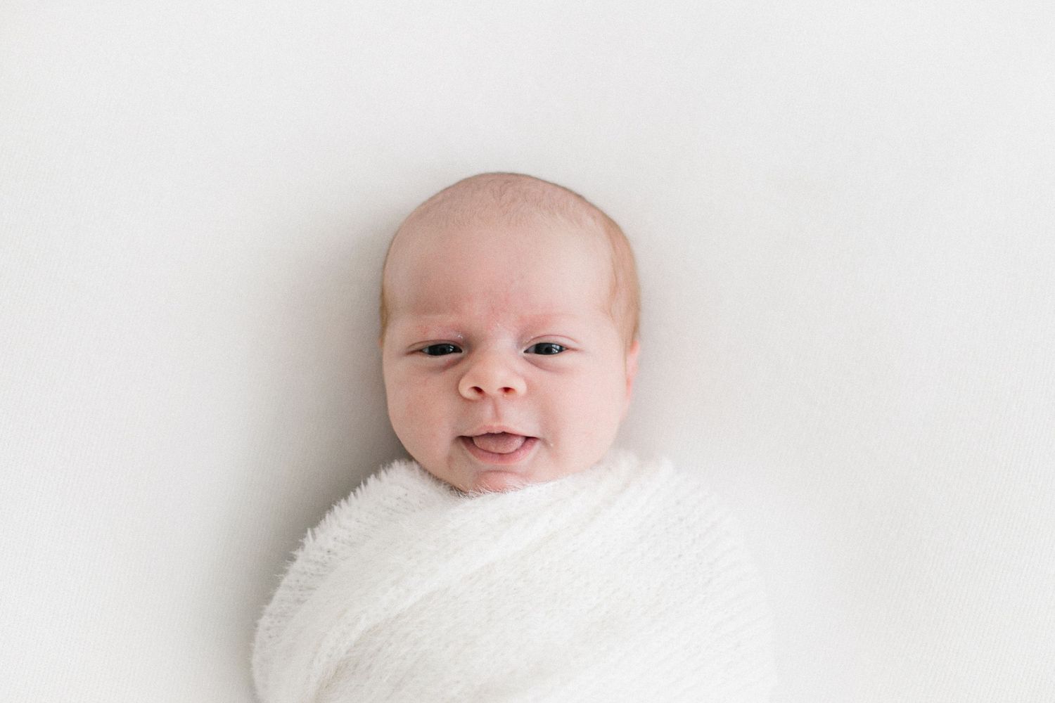 Smiling newborn baby swaddled in white wrap lying on white background.
