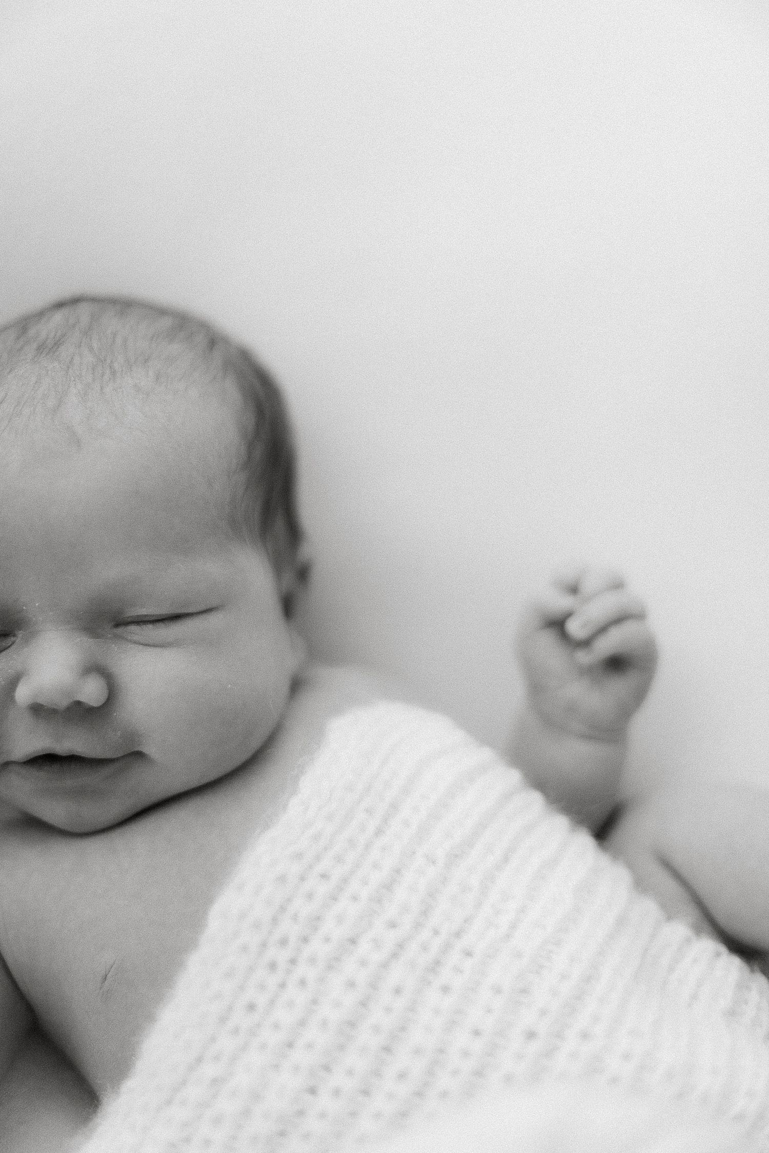 Peaceful sleeping newborn baby wrapped in a knit blanket with tiny fist raised, black and white photo.