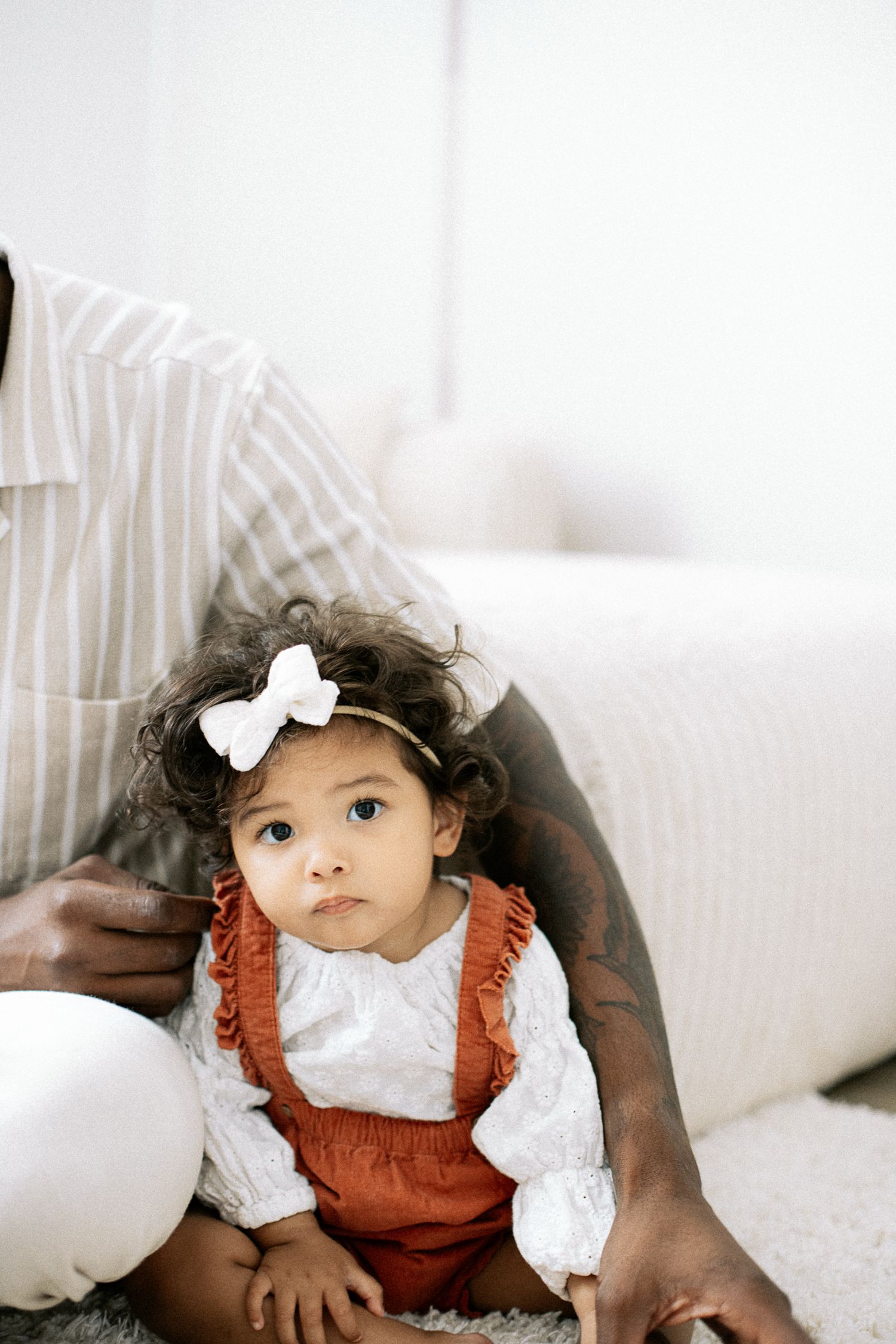 Adorable baby girl with curly hair and white bow headband wearing rust overalls sits on floor with parent.