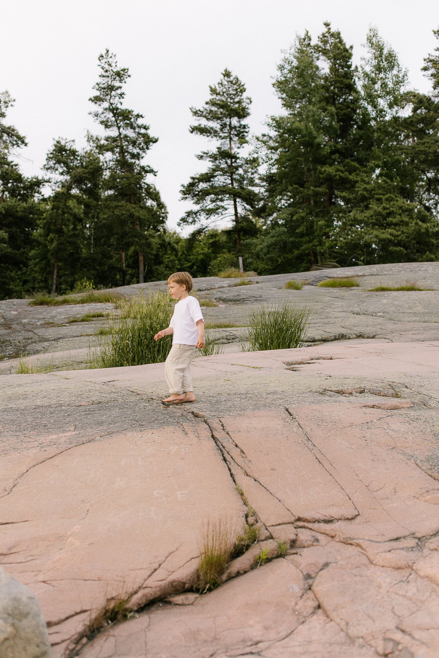 Young child walking barefoot on pink granite rocks with green trees in background.
