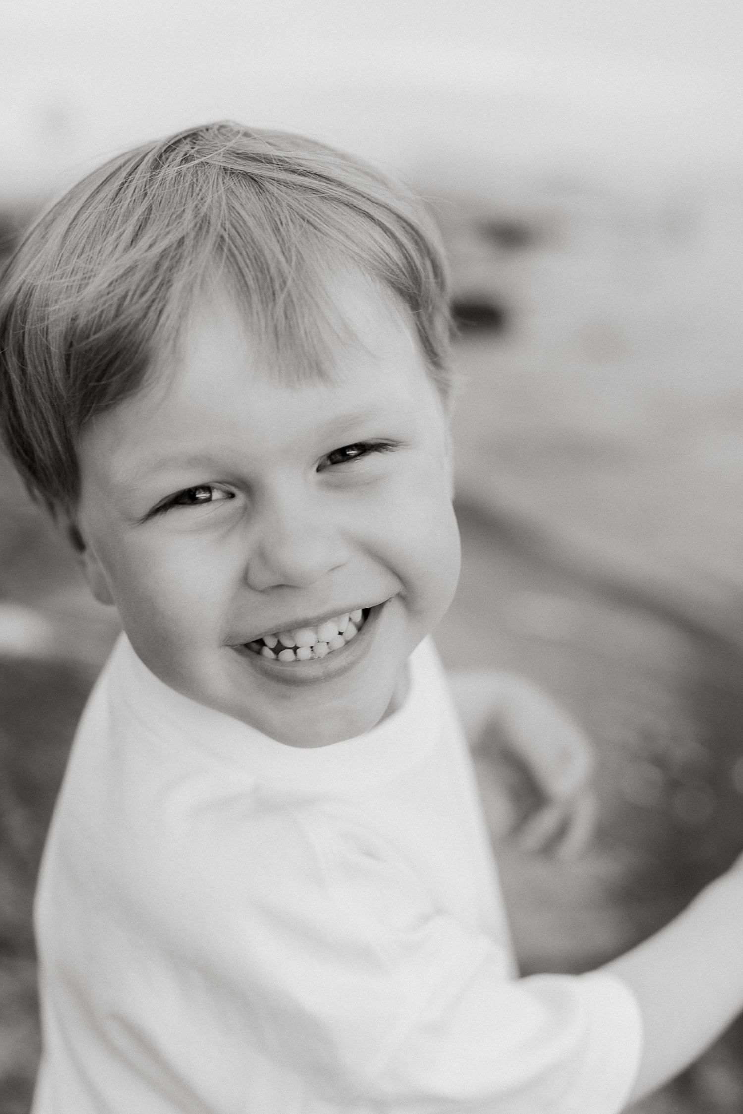 Black and white portrait of a smiling young boy with short hair wearing a white shirt outdoors.