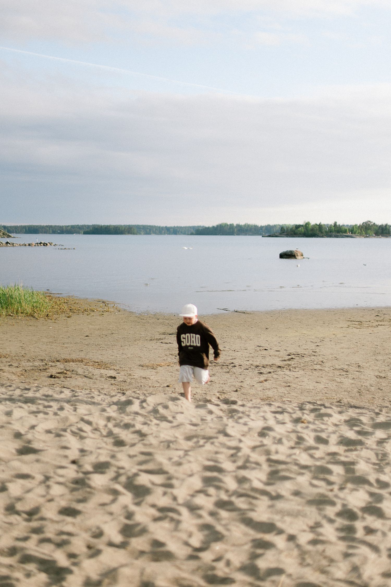 Child in black hoodie and white hat walking barefoot on sandy beach toward calm lake with tree-lined shore.
