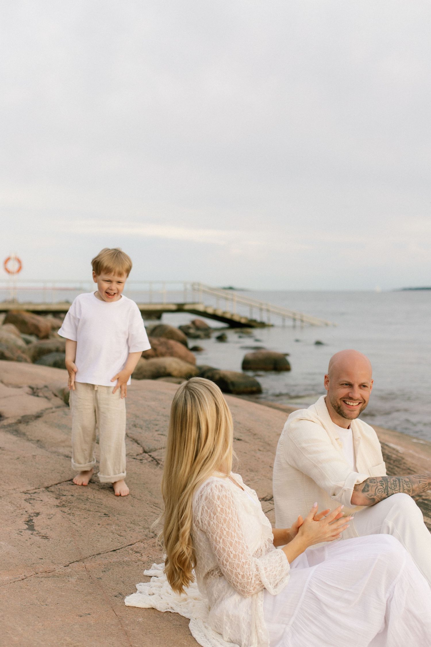 Happy family in white outfits relaxing by the rocky seaside, with a smiling boy and parents sitting near a pier.