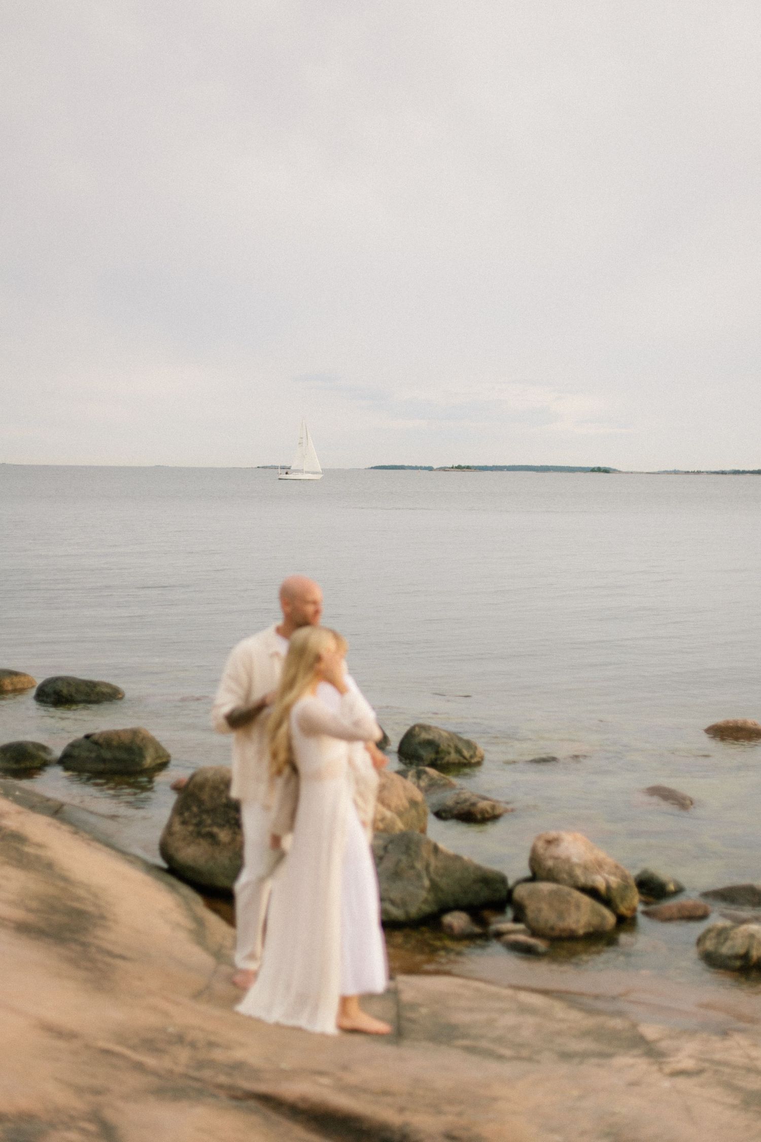 Couple in white embracing on rocky shore with sailboat on calm sea in background.