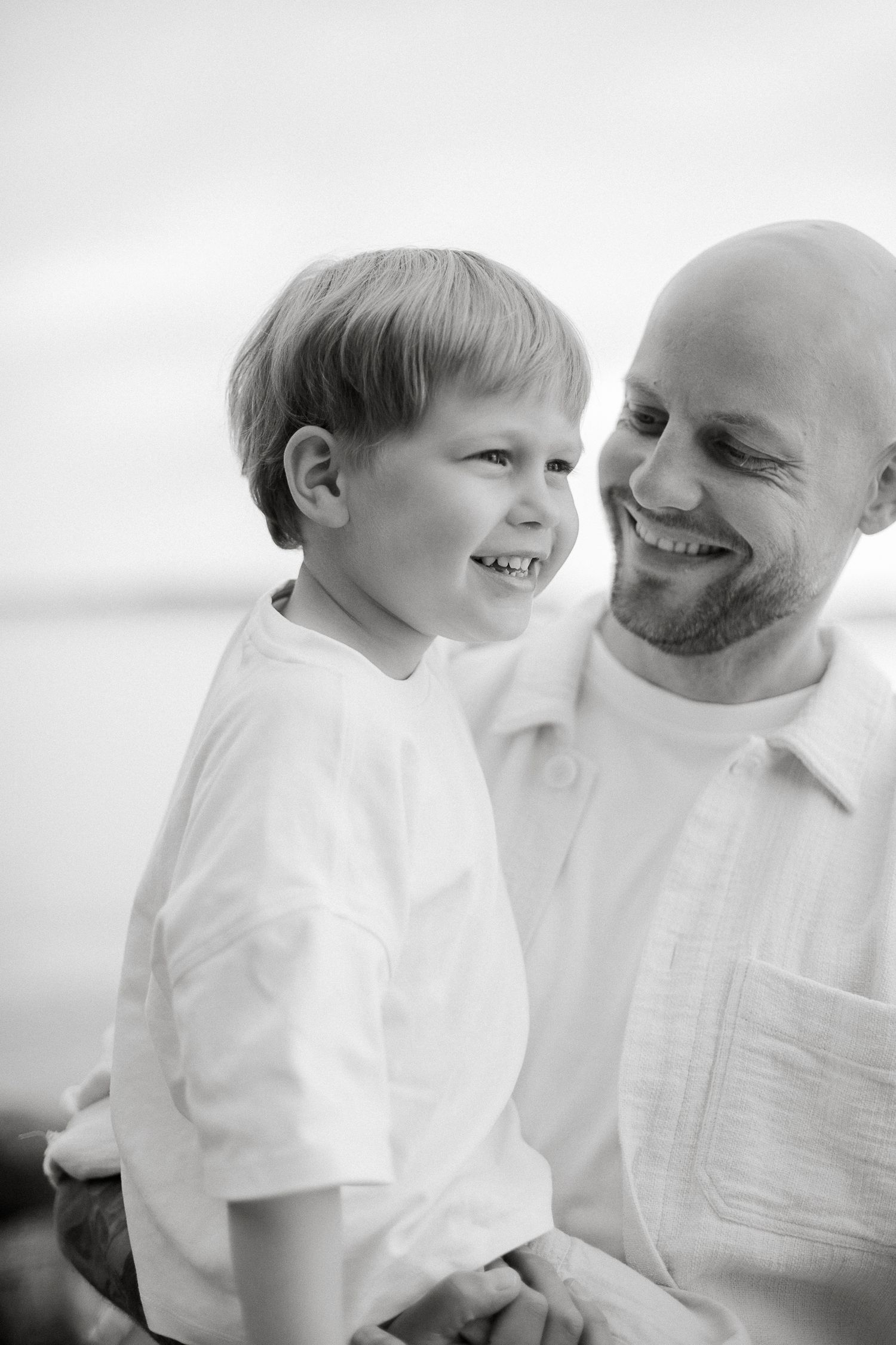Black and white photo of a smiling bald father holding his laughing young son outdoors near water.