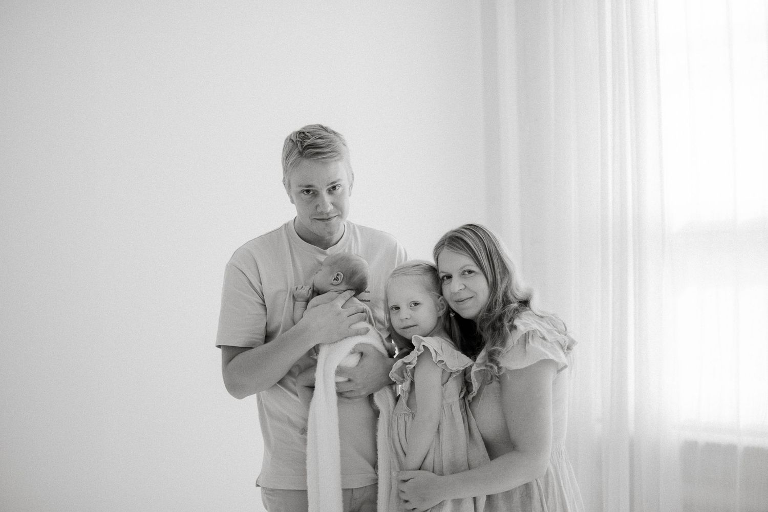 Black and white family portrait of parents with toddler and newborn baby near bright window.