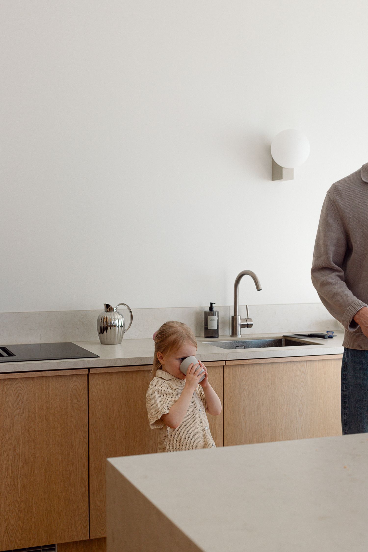 Toddler drinking from cup near kitchen sink beside adult in modern wooden kitchen.