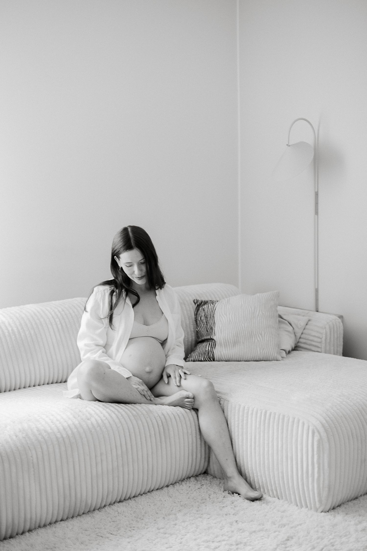 A pregnant woman sits barefoot on a couch, cradling her baby bump in a serene black and white photo.