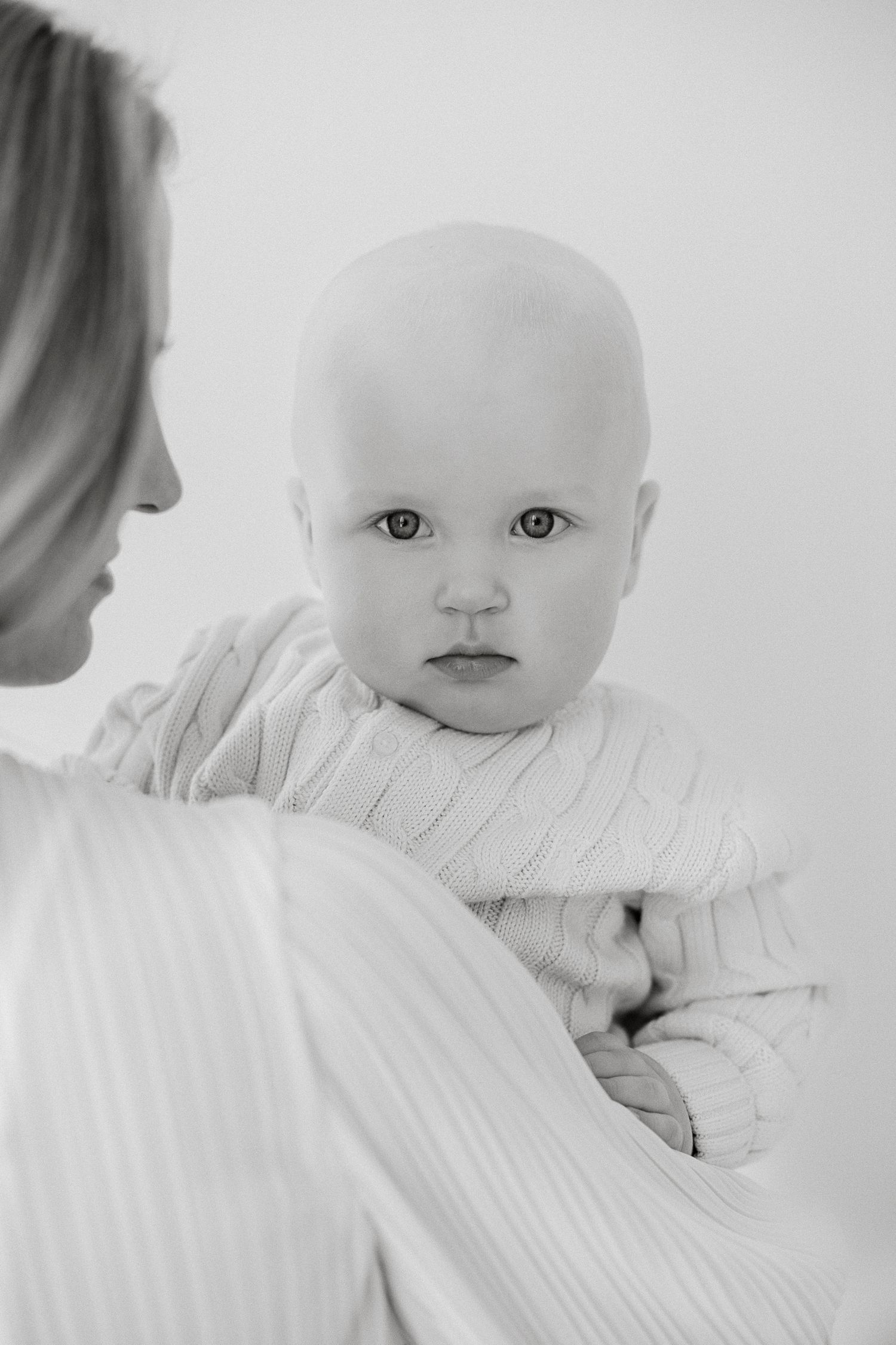 Black and white photo of a bald baby being held by a woman, gazing at the camera with curious eyes.