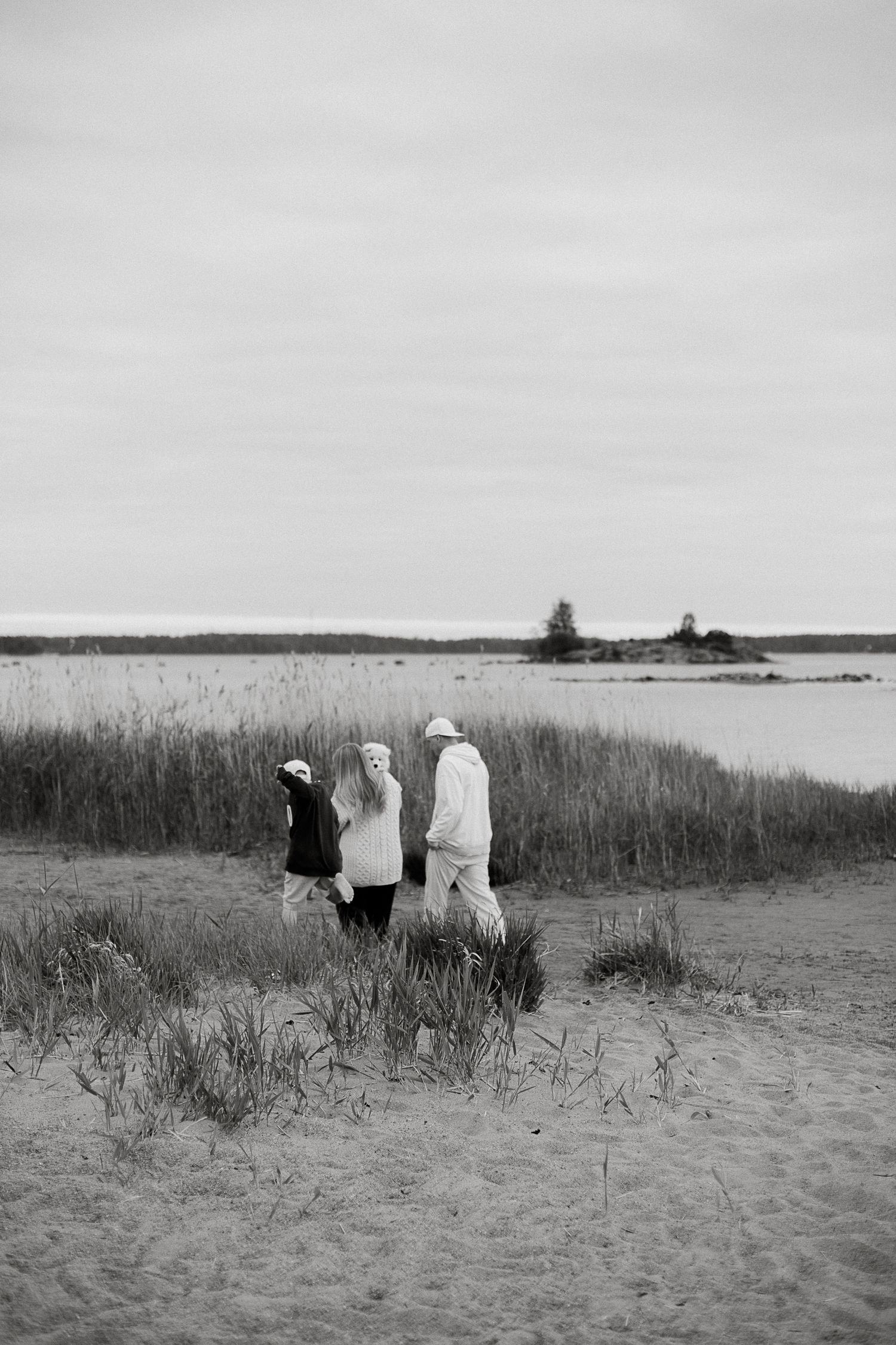 Black and white photo of three people walking on a sandy beach with tall grass and calm water in the background.