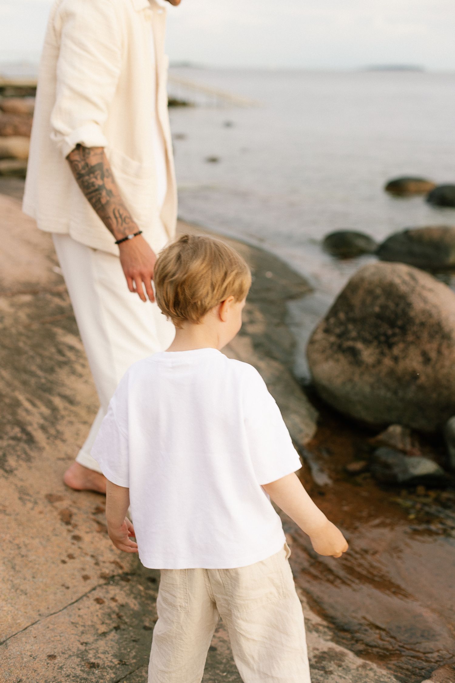 A tattooed parent and young child in white linen outfits walk barefoot on rocky shoreline near calm water.